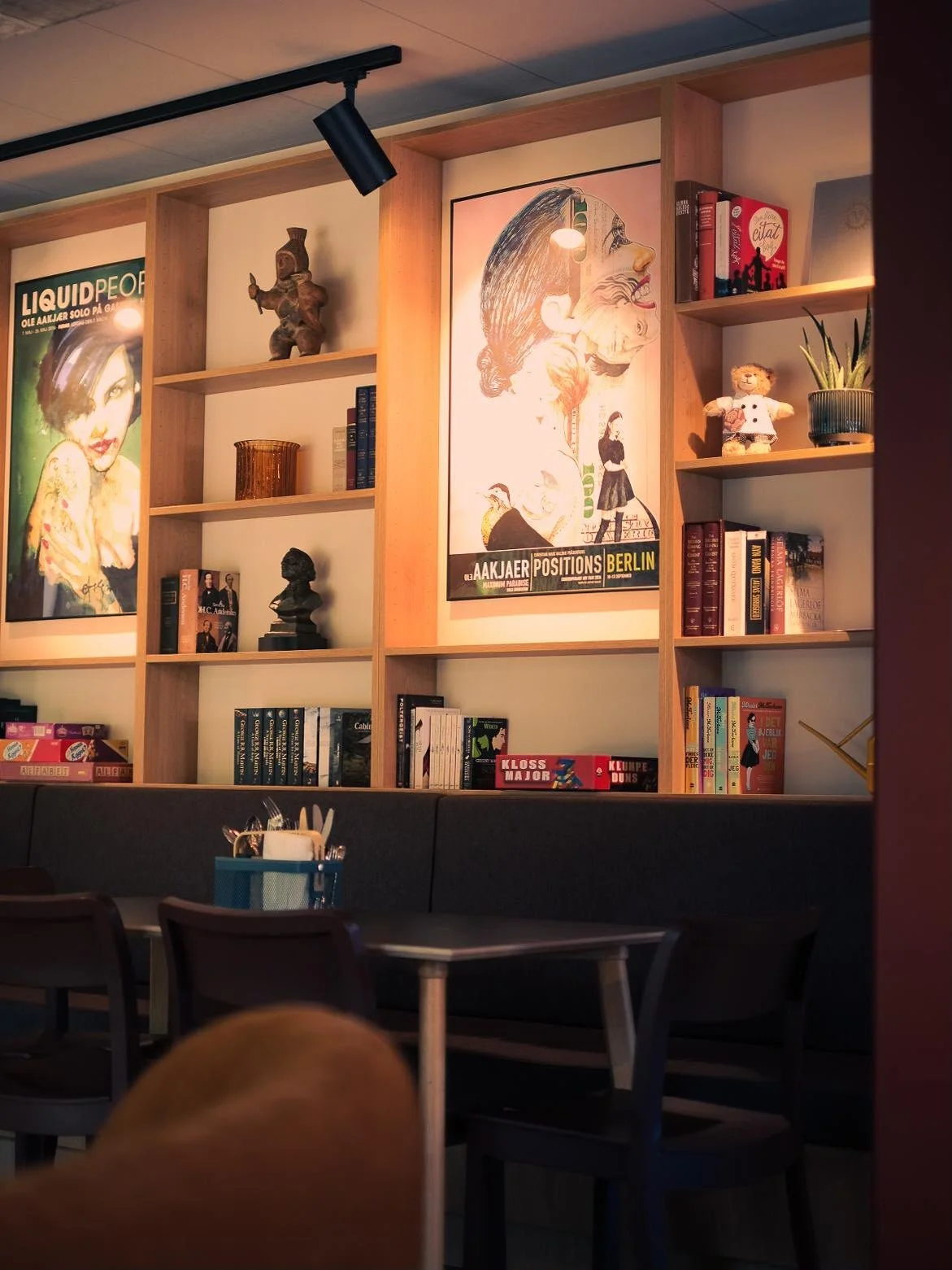Bookshelf with artwork, board games and decorative items at ALMA Cafe in Billund, including framed posters, books, and small sculptures.