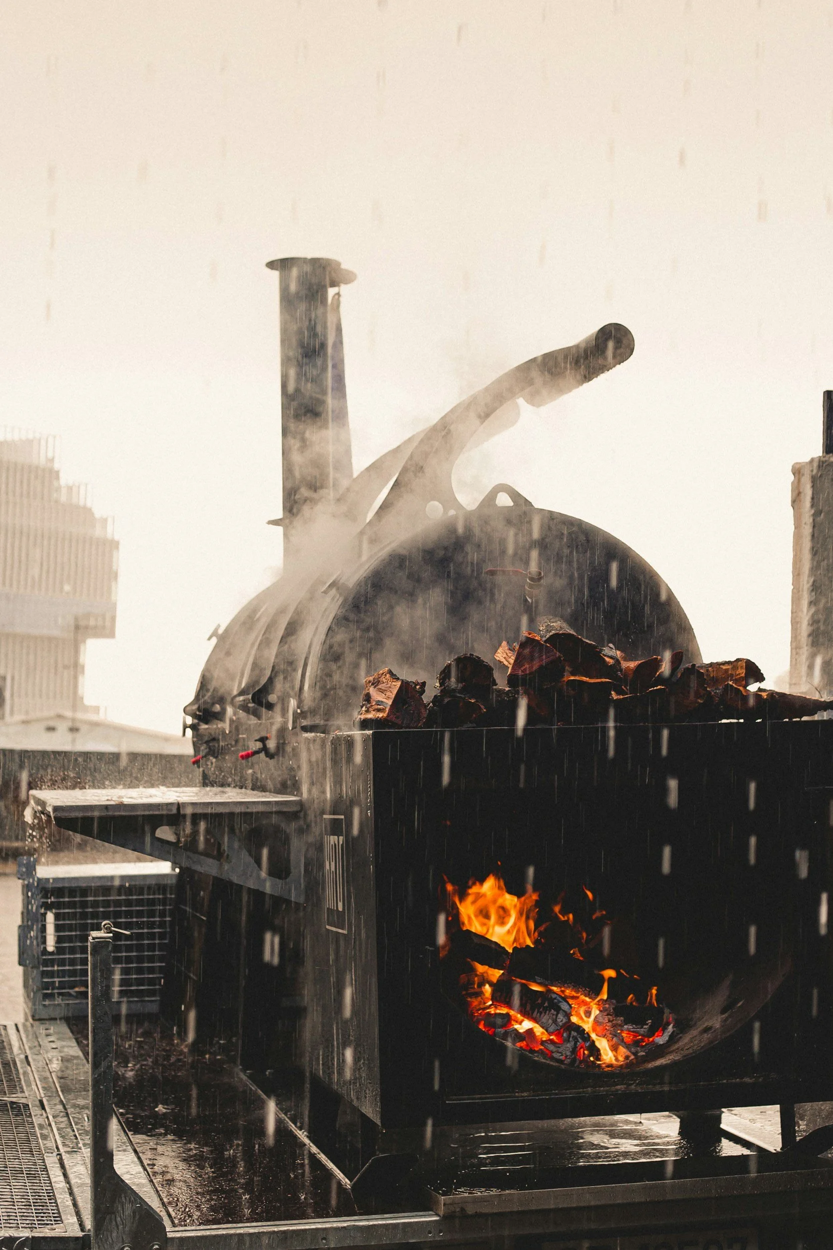 Industrial machine with open flame and burning coal, emitting smoke during a rainstorm in an urban setting.