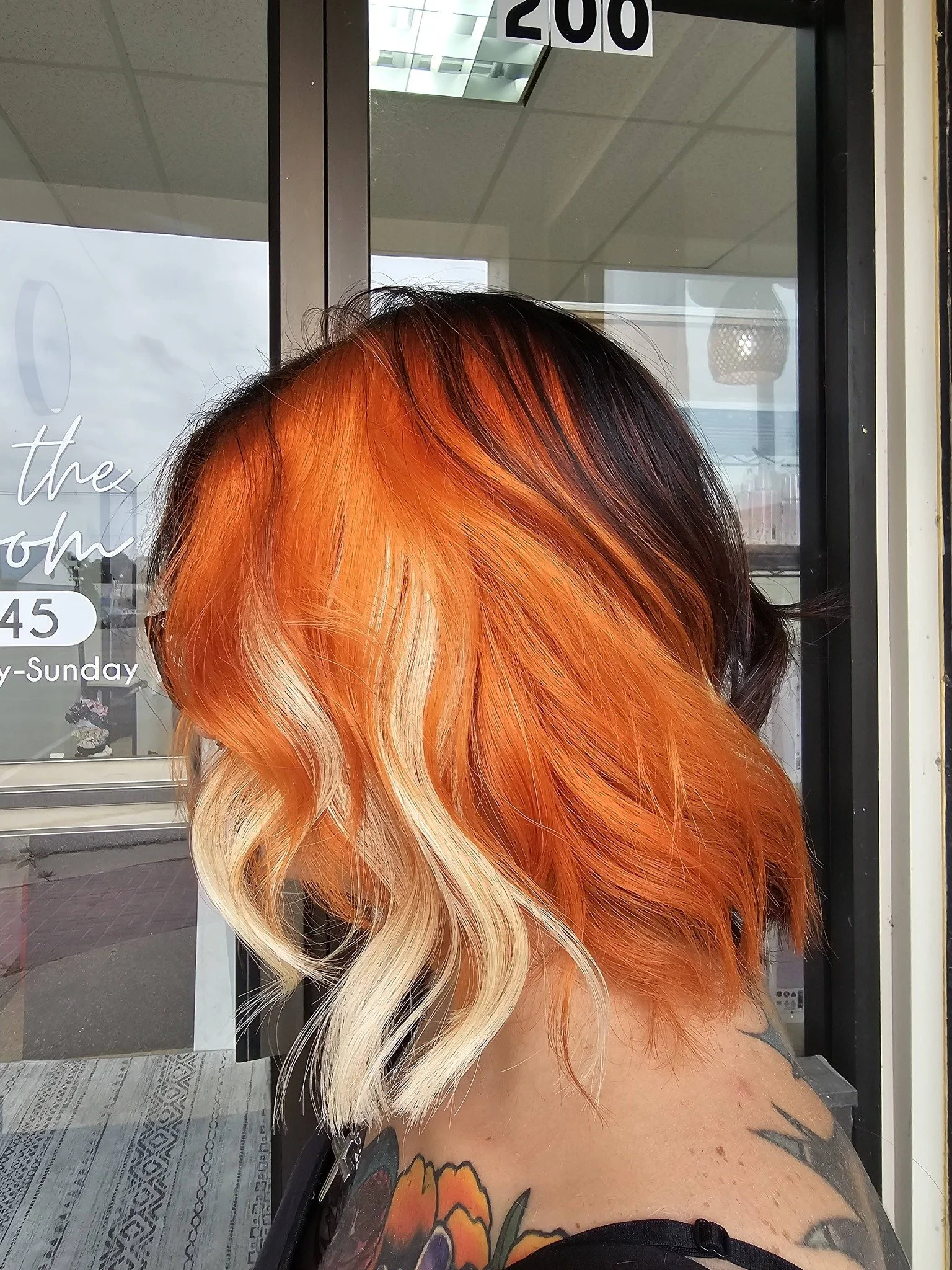 A person with vibrant, multi-colored hair featuring shades of orange, blonde, and dark brown. The person has tattoos on their shoulder and upper arm, and they are standing outdoors near a glass door with signage.