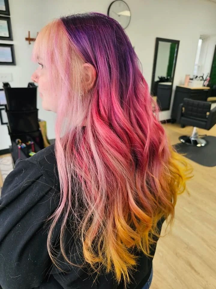 A woman with long, wavy hair dyed in multiple colors, including peach, pink, purple, and yellow, is sitting in a salon.