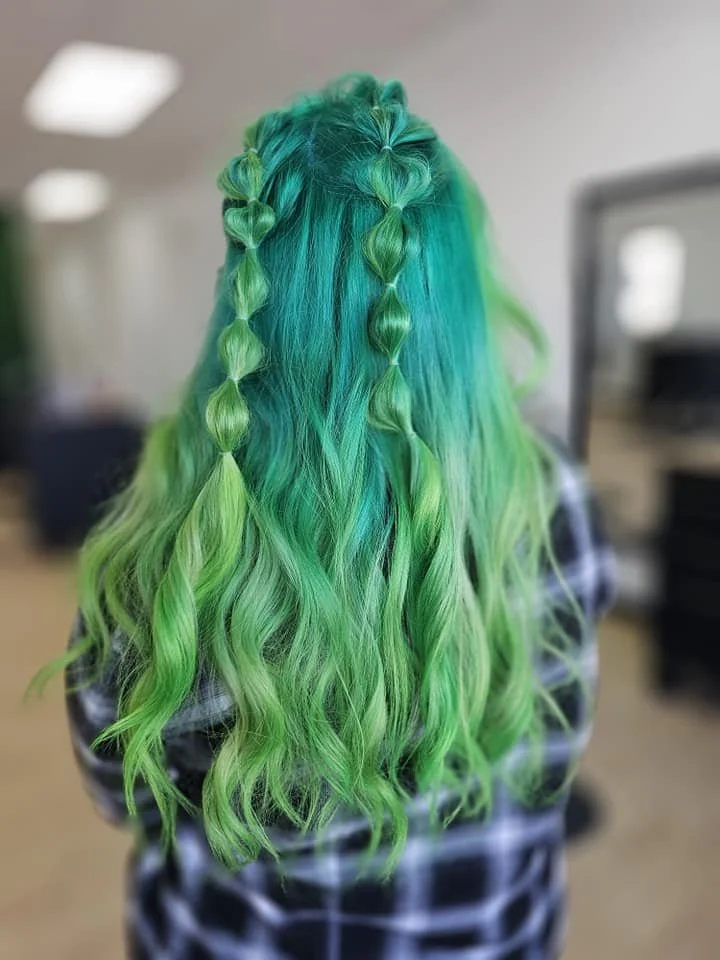 Back view of a person with long, wavy, bright green hair styled in heart-shaped braids on the top of the head.