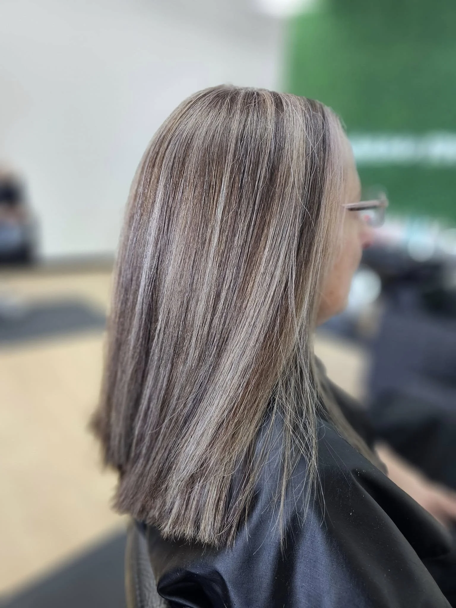 Close-up of a woman with long, straight, light brown hair, sitting in a salon chair with a black cape, and a blurred background featuring a green poster or wall.
