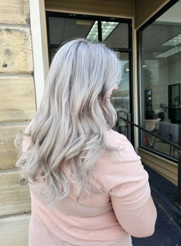 A woman with shoulder-length, wavy, silver-blonde hair standing outside near a building, wearing a pink top.