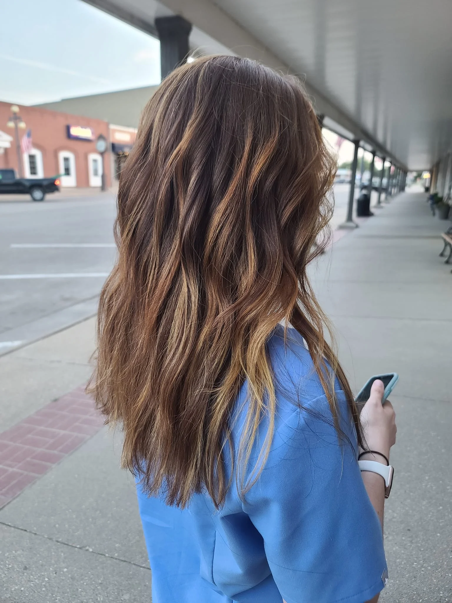 A woman with wavy, light brown hair stands outdoors, looking at her phone, wearing a blue top and a white watch on her left wrist, in an urban area with stores and street benches.