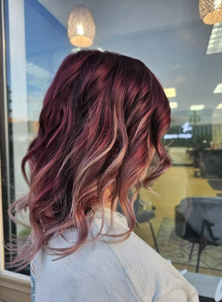Woman with wavy pink and brown ombre hair, seen from the side in front of a window with indoor and outdoor reflections.