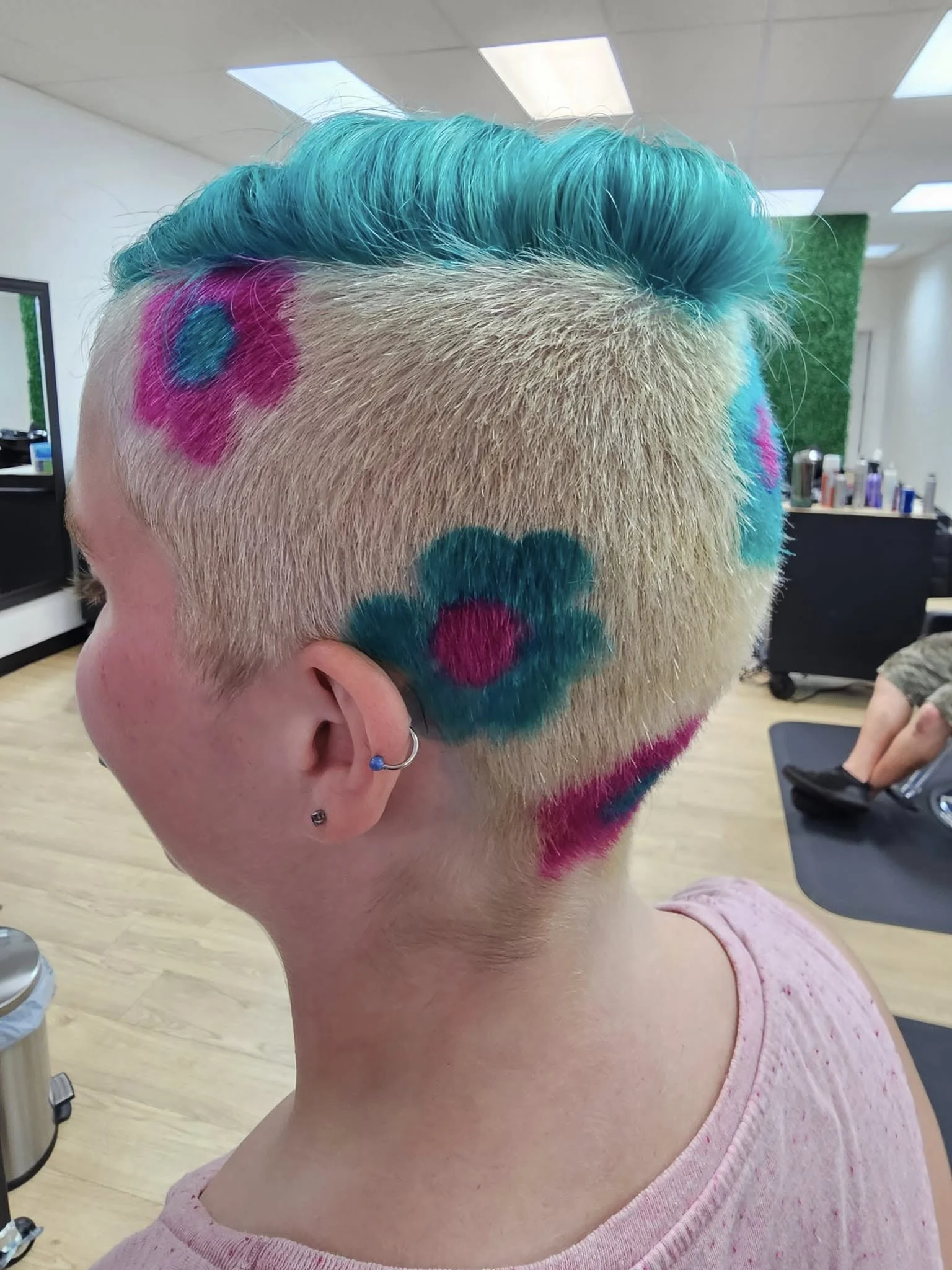 A person with short blonde hair featuring a colored pattern of pink, teal, and purple dots and streaks, with the top section dyed teal and styled voluminously.
