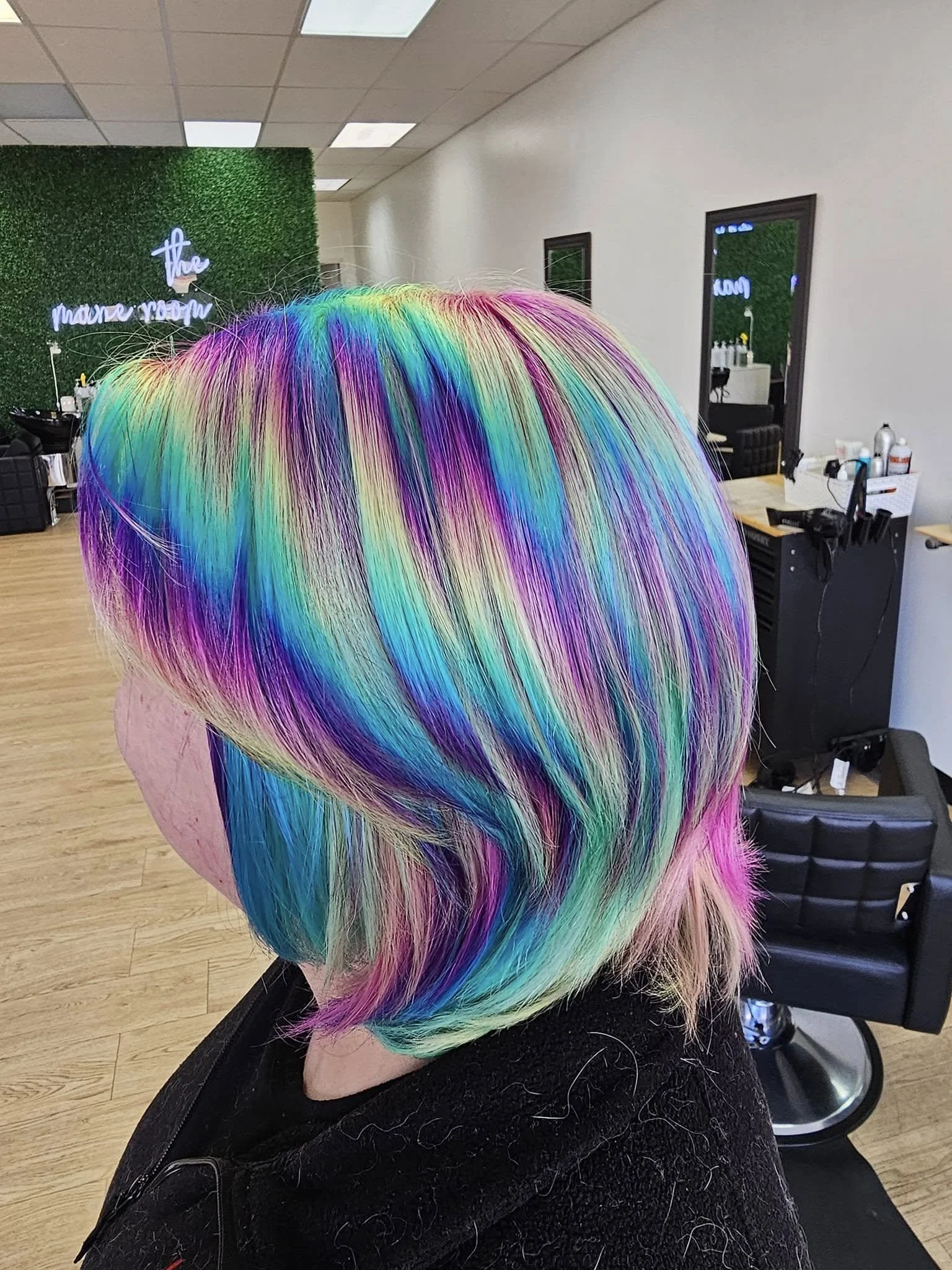 A person's head with rainbow-colored, vibrant, multi-colored hair styled in a bob cut, inside a hair salon with salon chairs, mirrors, and hair products visible in the background.