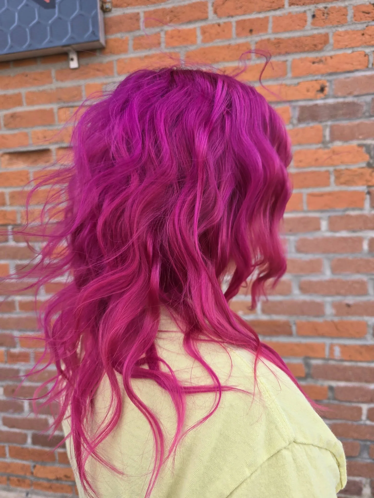 Person with long, wavy, vibrant magenta hair wearing a light yellow top in front of a brick wall.
