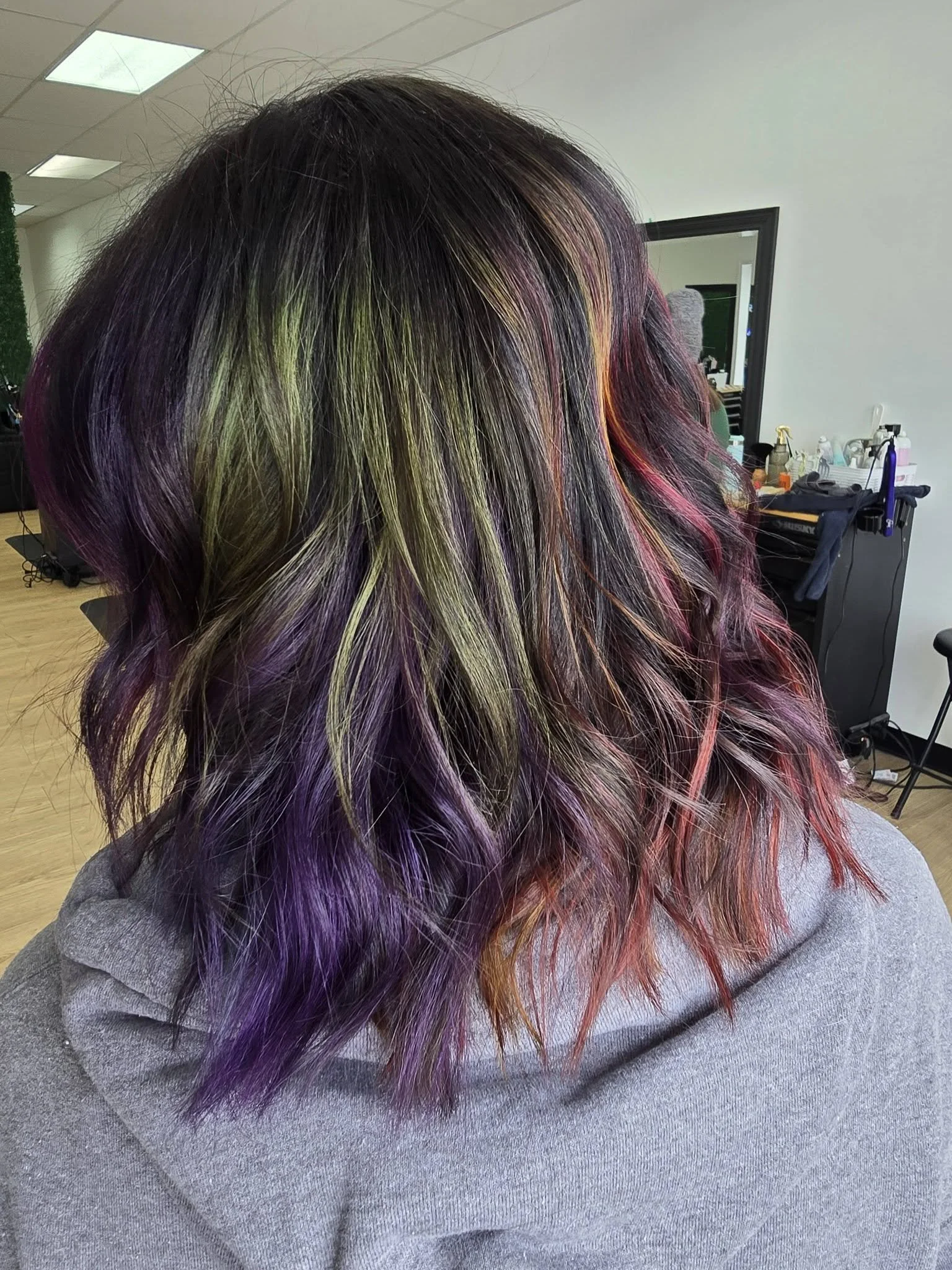 Woman with shoulder-length hair dyed in multiple vibrant colors including green, purple, red, and orange, sitting in a salon or studio with a mirror, equipment, and supplies in the background.