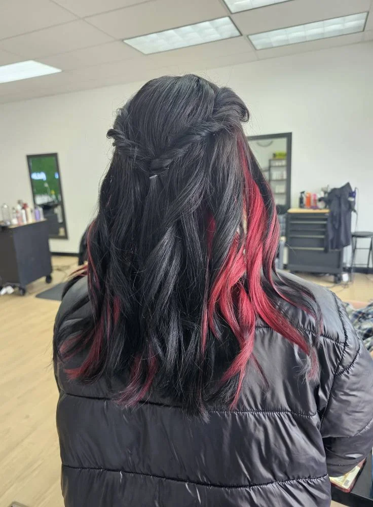 Back view of a woman with black hair styled with a braid crown on top and pink highlights in her wavy hair, wearing a black puffy jacket, inside a salon.