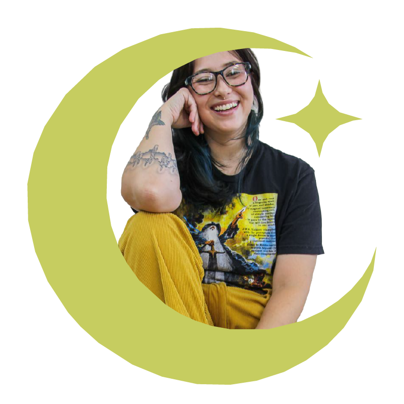 Young woman with glasses and tattoos smiling, wearing a graphic t-shirt and yellow pants, sitting and resting her head on her hand.