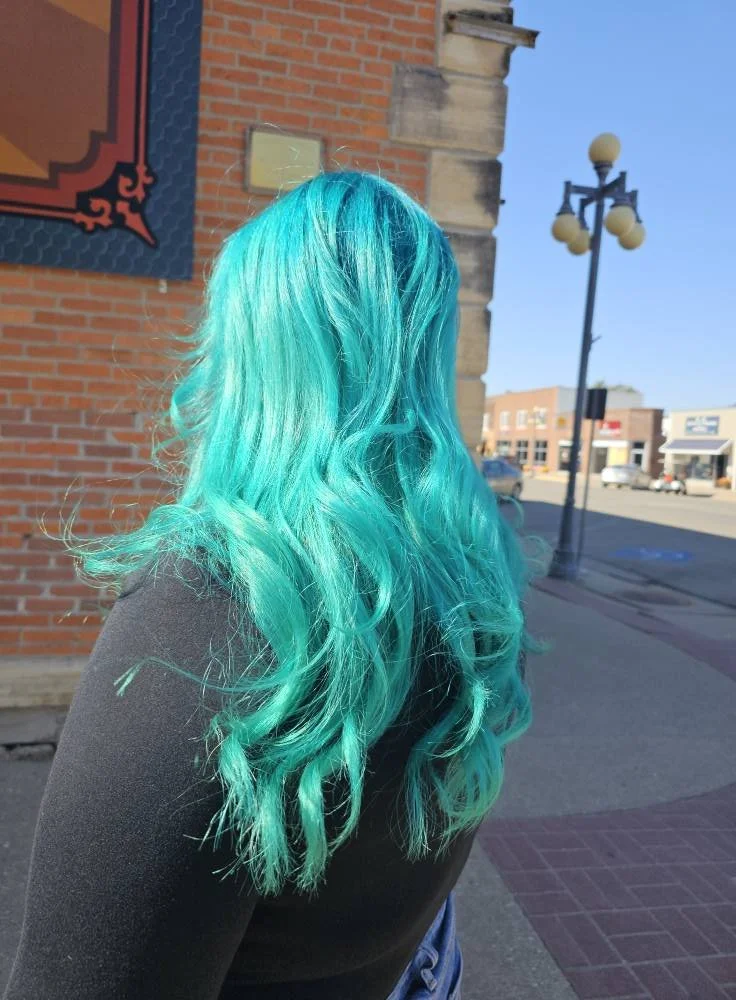 Back view of a woman with long, wavy, bright turquoise hair standing on a city sidewalk with brick and stone buildings and a streetlamp in the background.