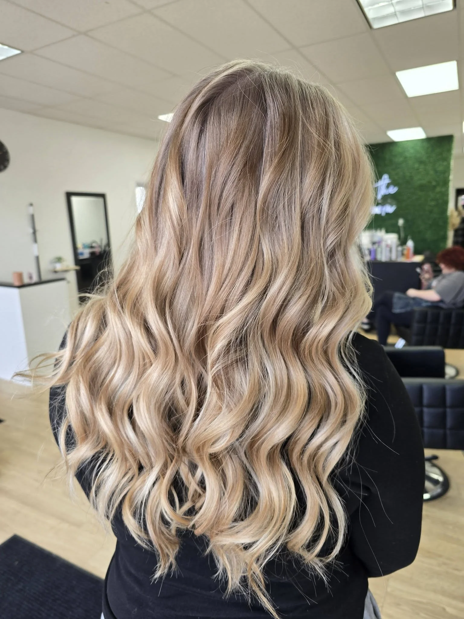 Long wavy blonde hair with darker roots, shown from the back in a salon or studio setting.
