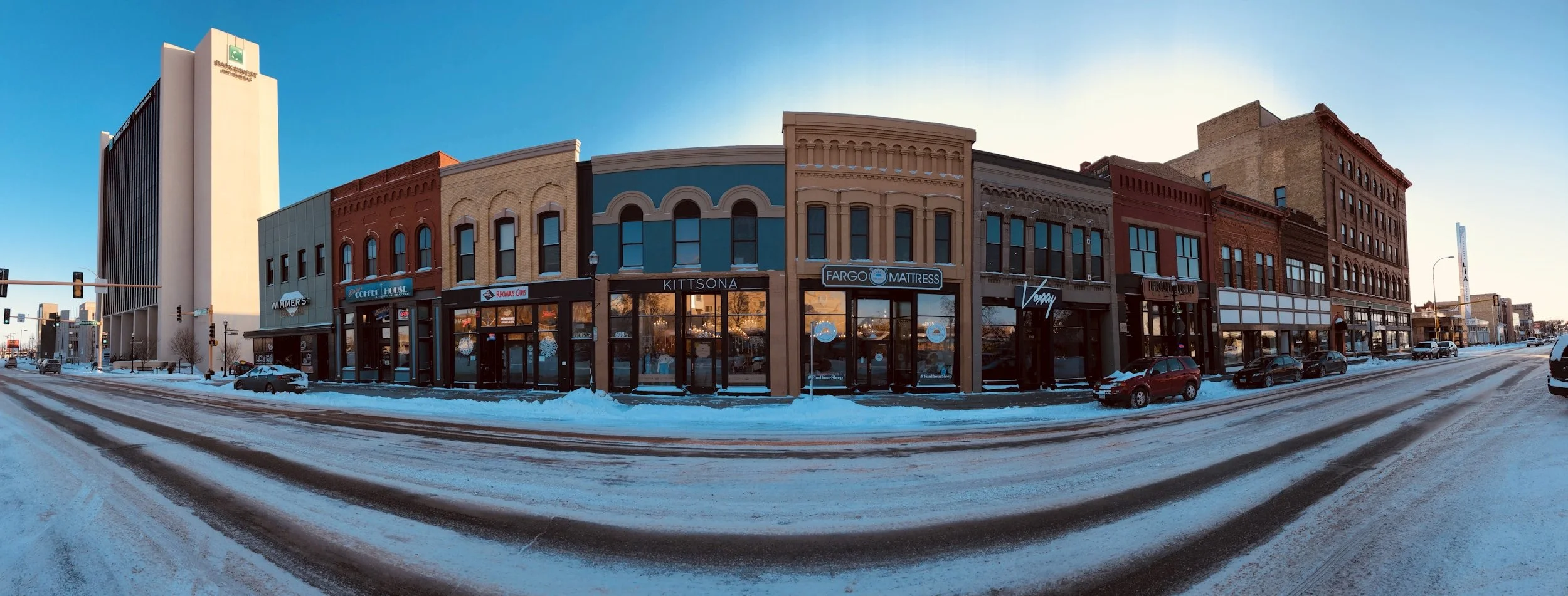 A street view of a downtown area with shops and buildings, snow on the ground, and cars parked along the street during sunset or sunrise.