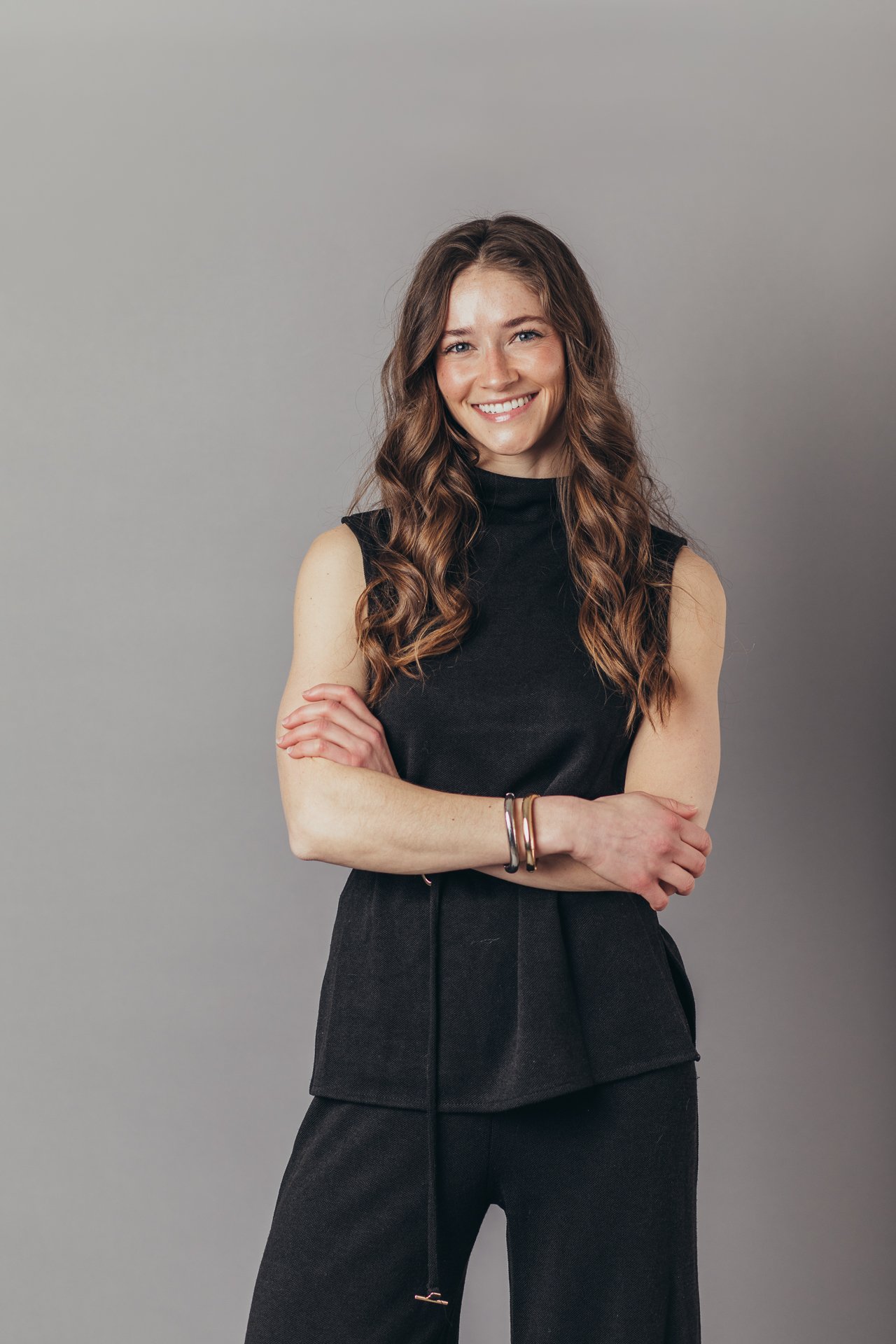 Young woman with long, wavy brown hair smiling, wearing a black sleeveless top and dark wide-leg pants, standing against a plain gray background.