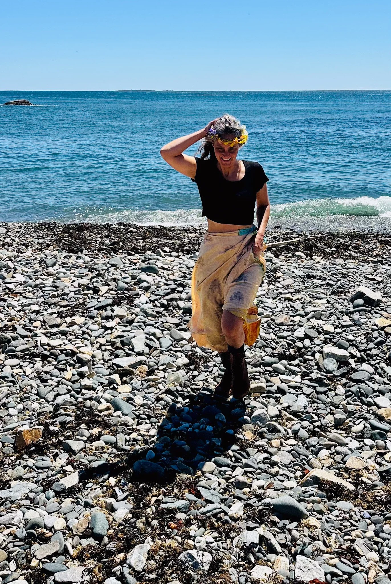 Woman with a flower crown smiling and walking on a rocky beach near the ocean.