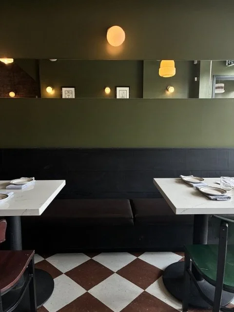 Interior of a restaurant with two tables set with napkins and plates, a dark bench seat, and a mirror on the wall reflecting other parts of the restaurant, decorated with warm lighting and framed pictures.