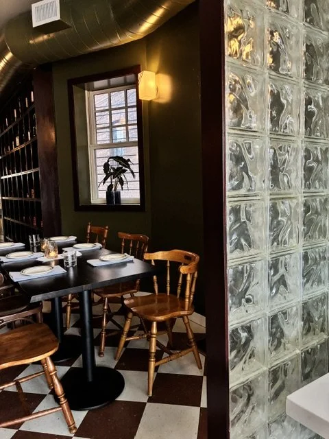 A dining area in a restaurant with a black table set for four, surrounded by wooden chairs. There is a window with a plant on the windowsill, a wall-mounted light fixture, and a glass block wall dividing the space. The floor has a black and white che