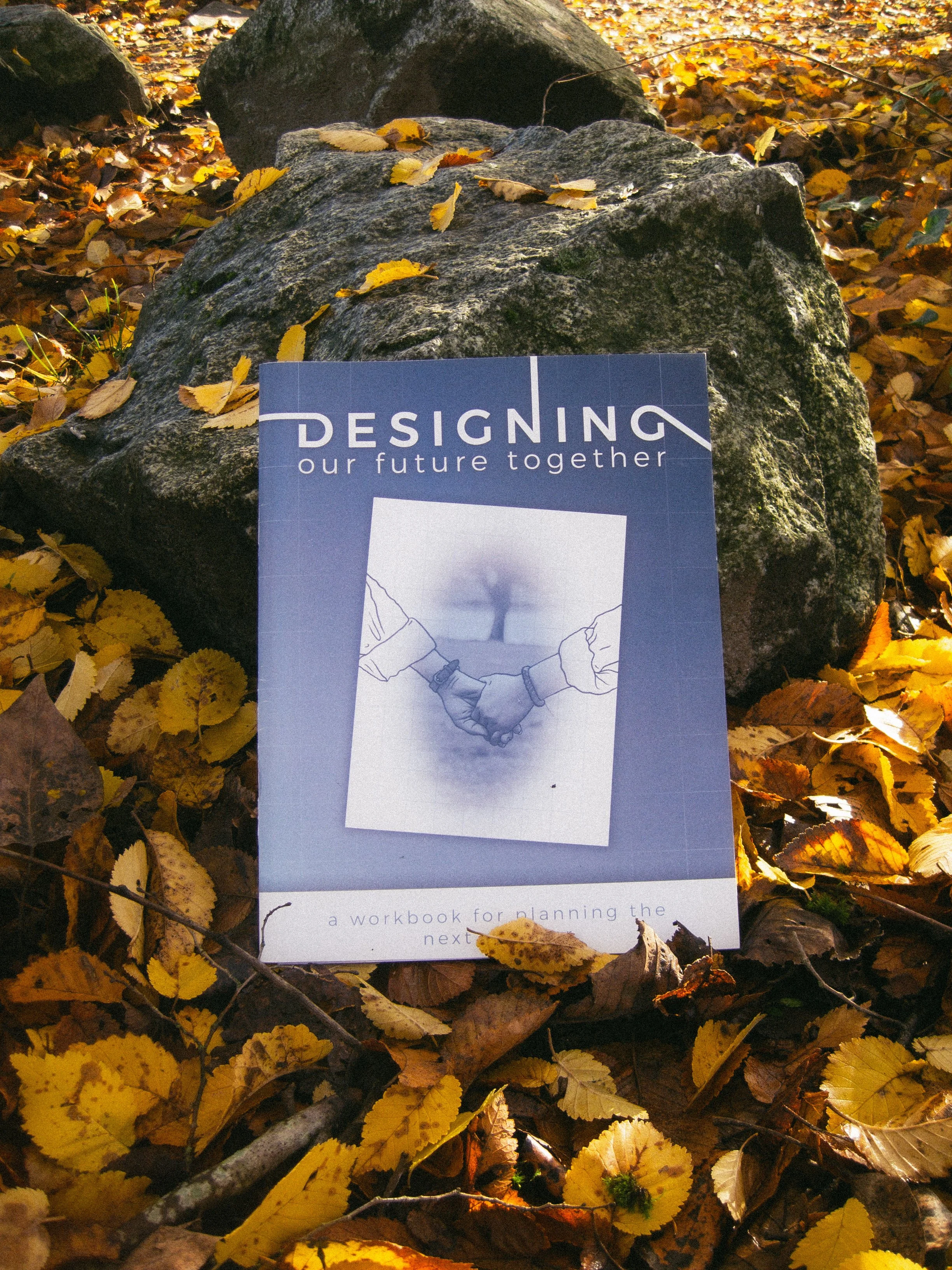 A book titled 'Designing our future together' lies among fallen yellow and brown autumn leaves on the ground, with rocks around it.