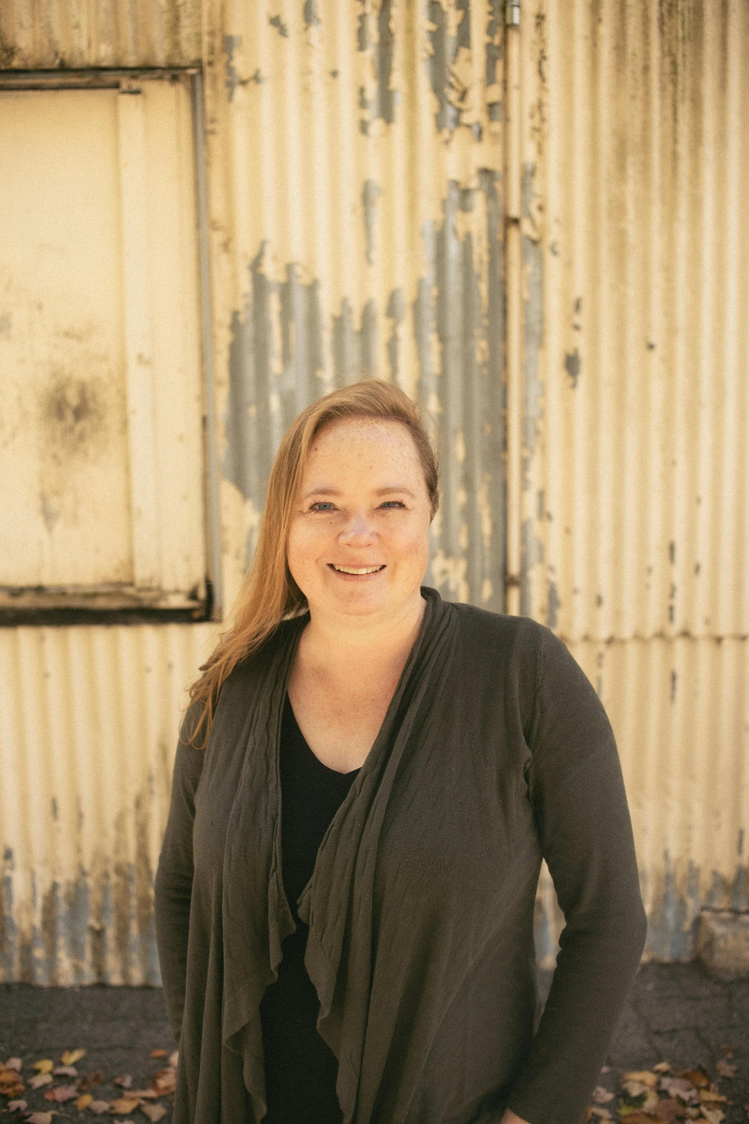 A woman with long hair, wearing a black shirt and jacket, stands outdoors in front of a weathered corrugated metal wall with peeling paint, fallen leaves on the ground, and a window to her left.