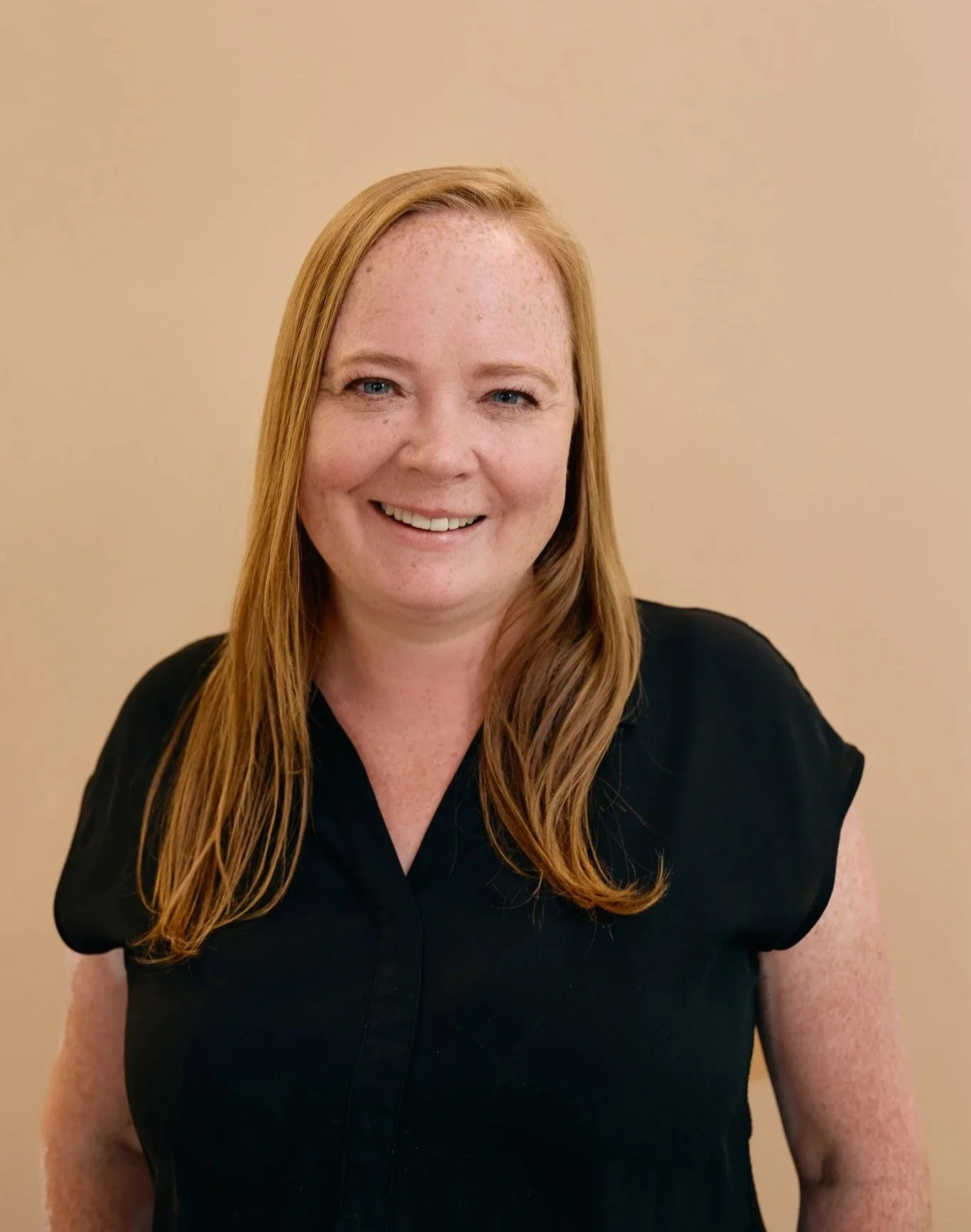 A woman with red hair and freckles smiling, wearing a black top, in front of a beige wall.