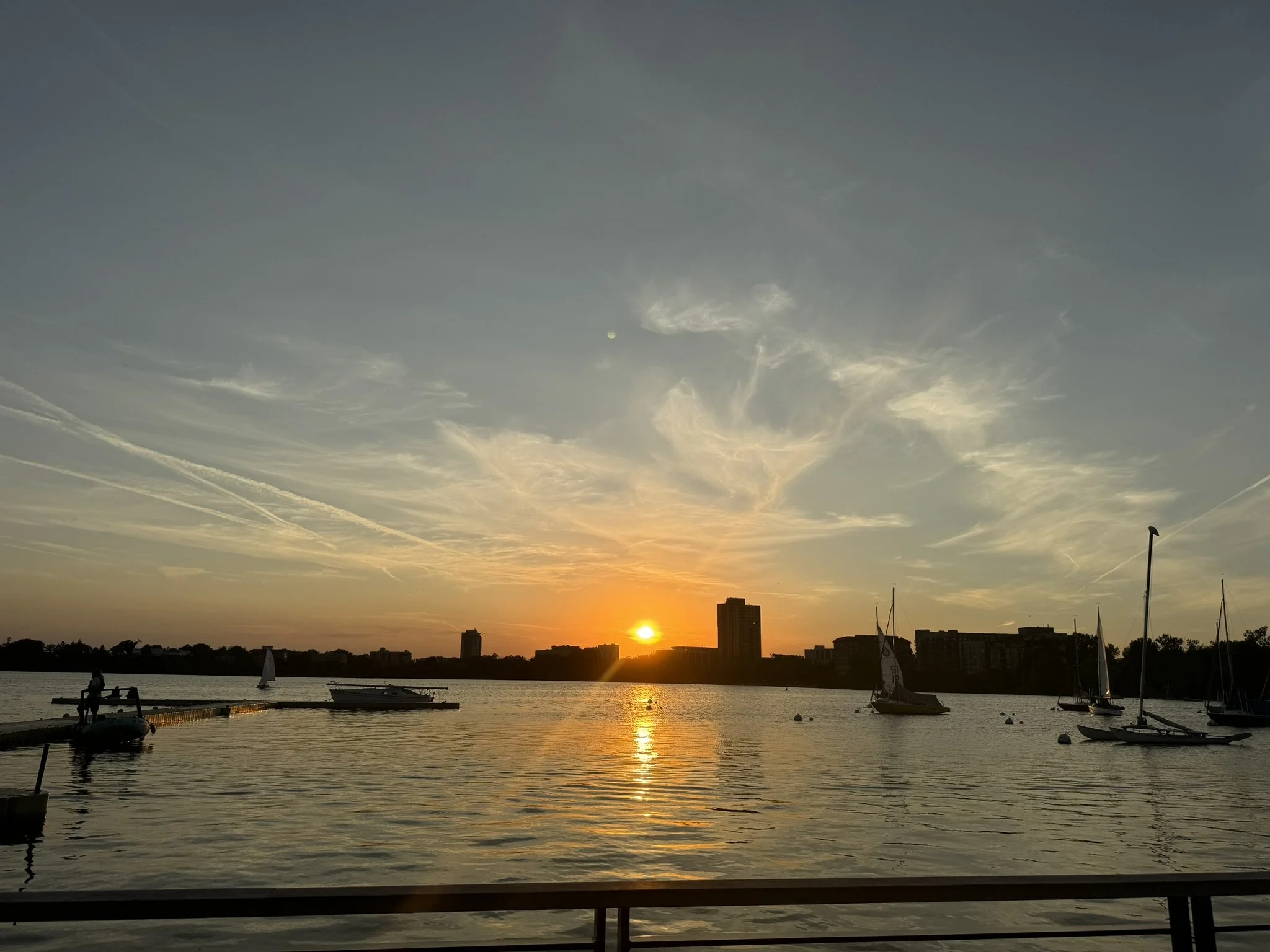 Sun setting over a lake with sailboats, a dock, and a city skyline in the background.