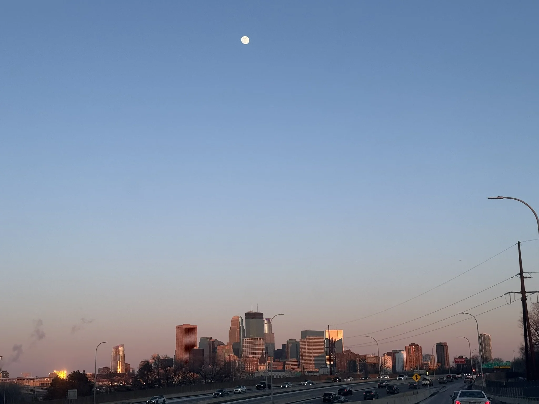 City skyline with high-rise buildings during sunset, with a clear blue sky and a visible moon.