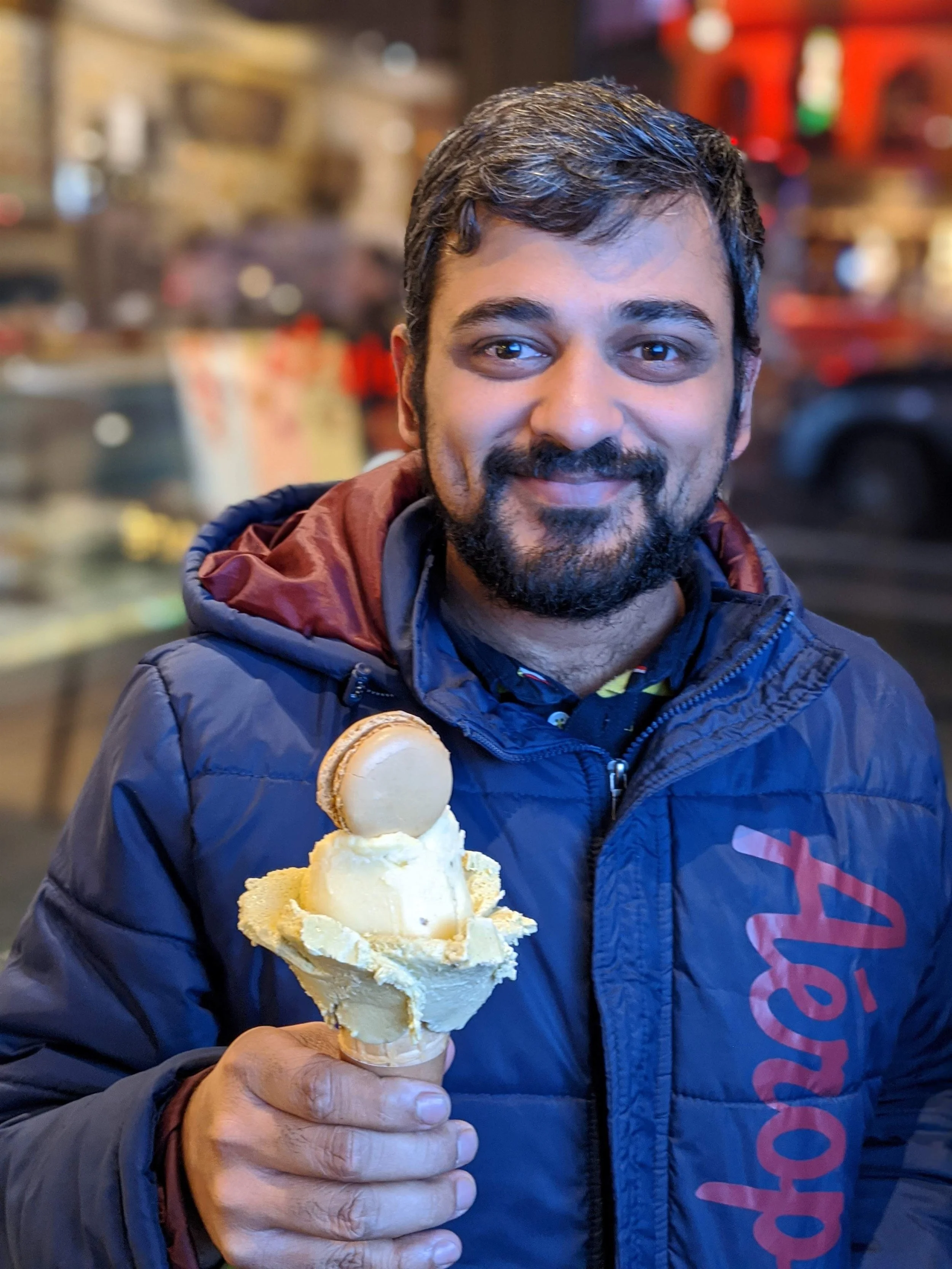 A smiling man with dark hair and a beard holding a large ice cream cone with a scoop of vanilla ice cream and a cookie on top inside a restaurant or cafe with a blurred background of shelves and decor.