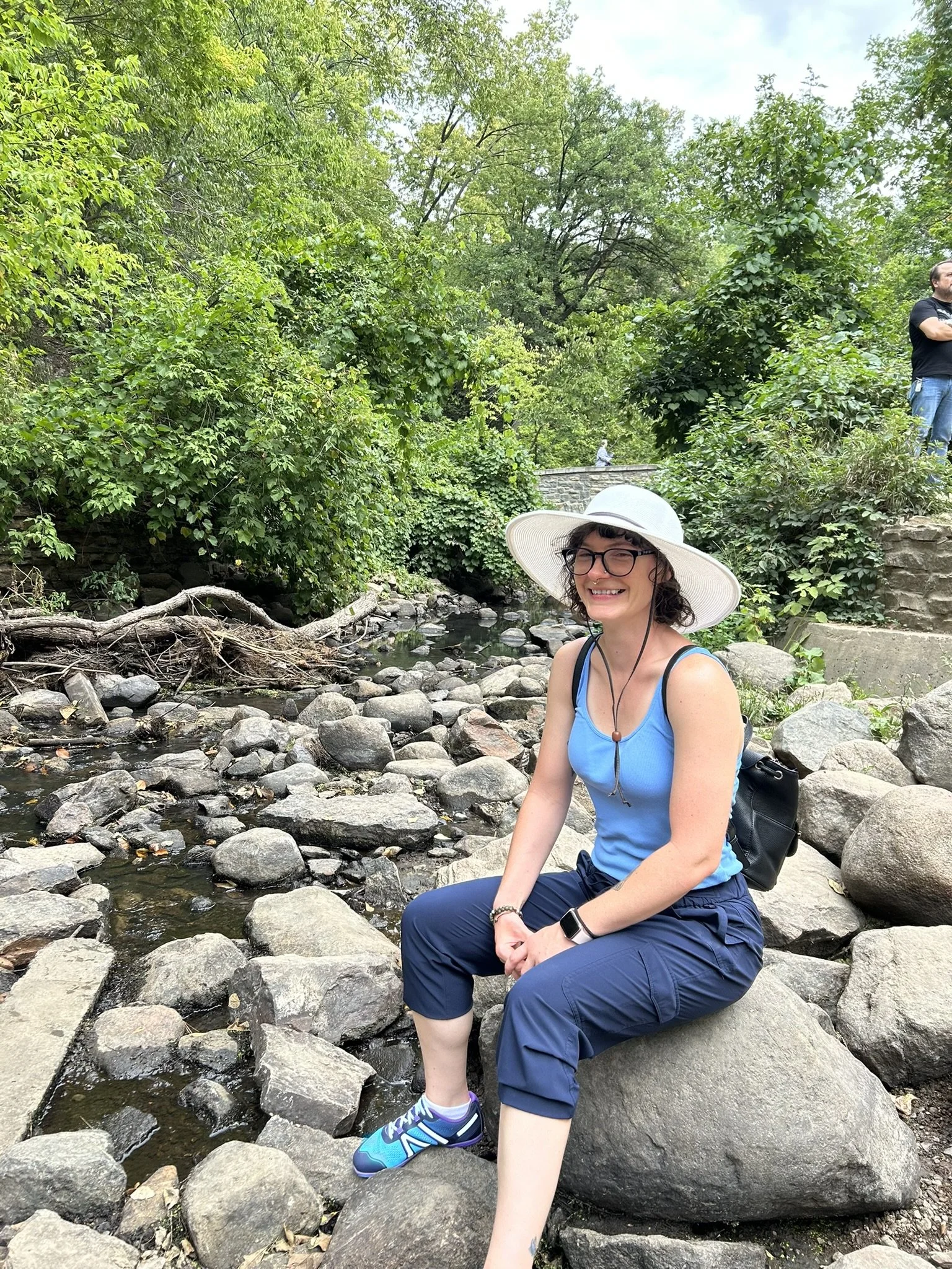 A woman sitting on rocks near a small creek, smiling and wearing a wide-brimmed white hat, glasses, and a blue tank top, in a lush green forest setting