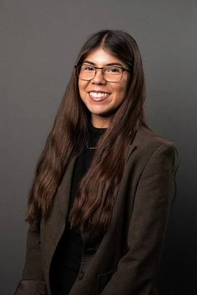 A woman with long dark hair, glasses, and a brown blazer smiling against a gray background.