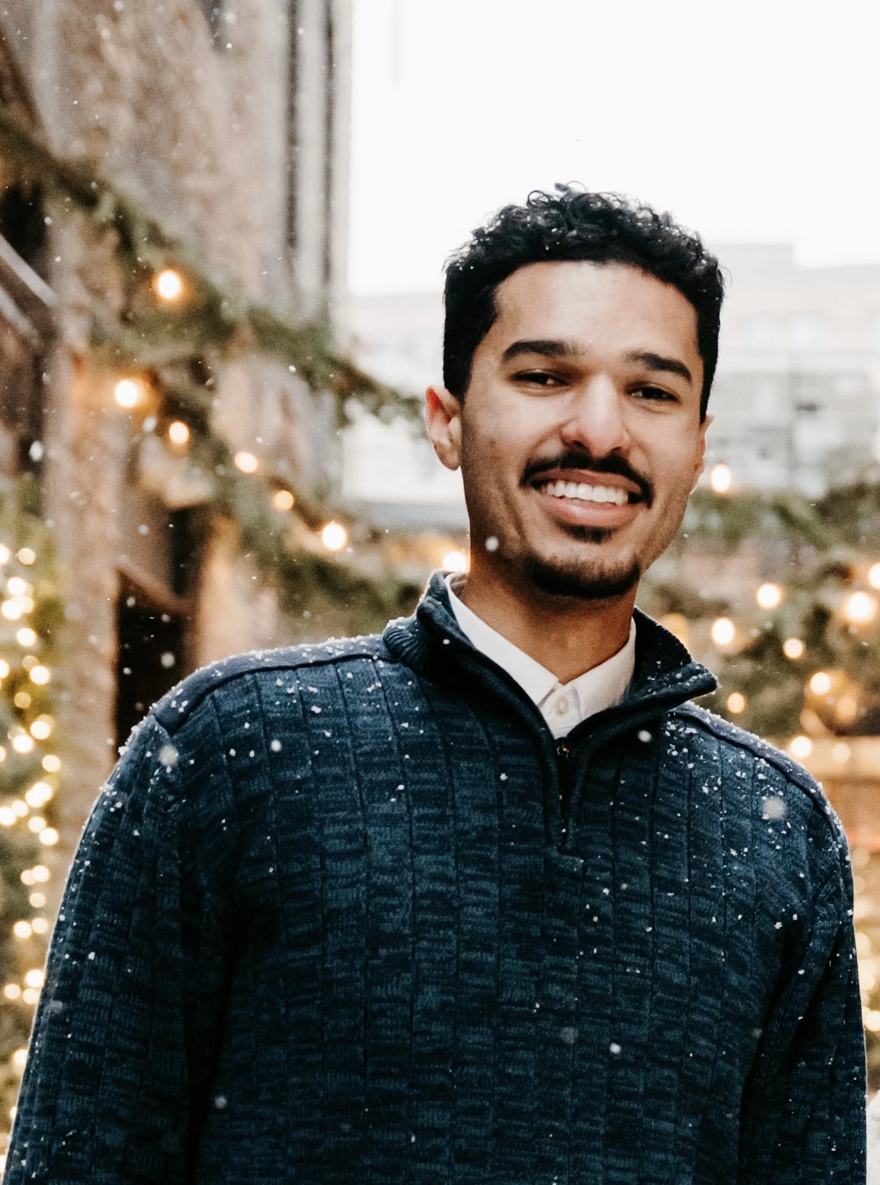 A young man with dark curly hair, dark facial hair, and light brown skin smiling outdoors during snowy weather, with decorated Christmas trees and lights in the background.