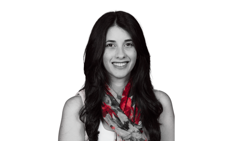 Black and white portrait of a smiling woman with long dark hair, wearing a light-colored top and a patterned scarf with red and gray accents against a solid black background.