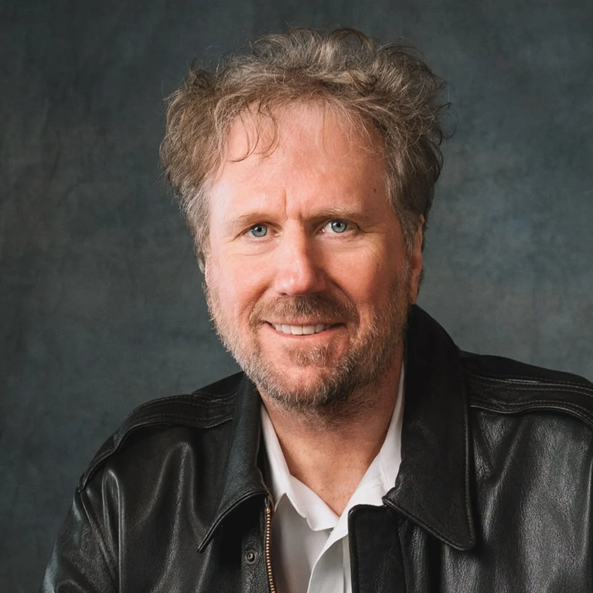 Portrait of a middle-aged man with light brown hair, blue eyes, a beard, and mustache, wearing a white shirt and black leather jacket, smiling against a dark gray background.