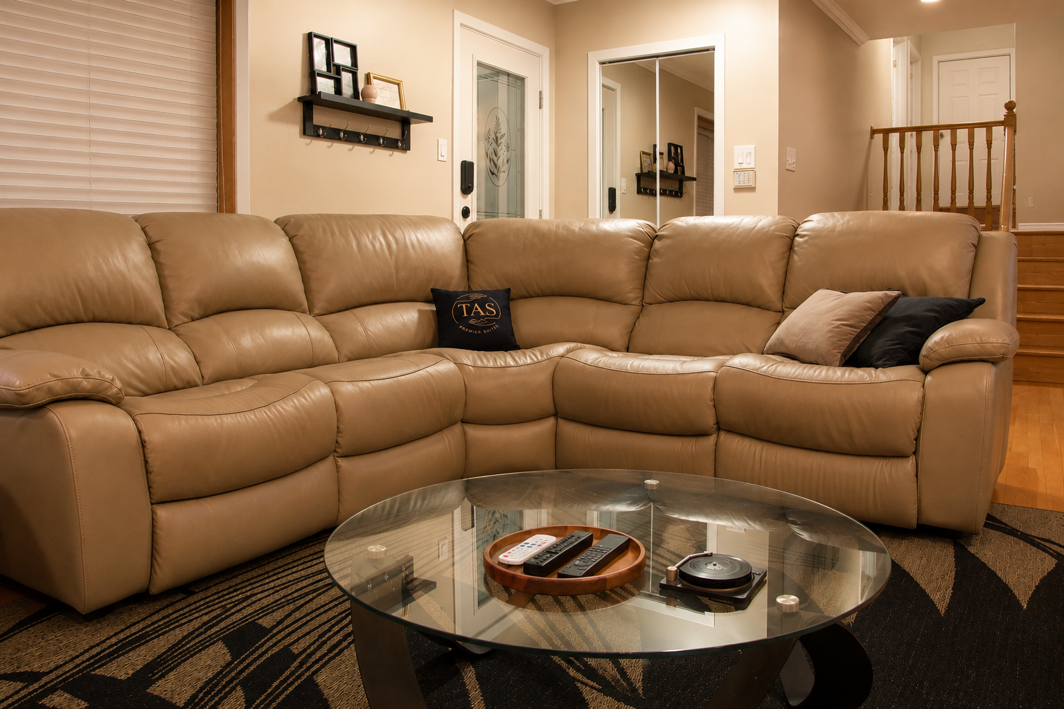 Living room with a large tan leather sectional sofa, glass coffee table with remotes and a robotic vacuum, and a wall with picture frames and a mirror.