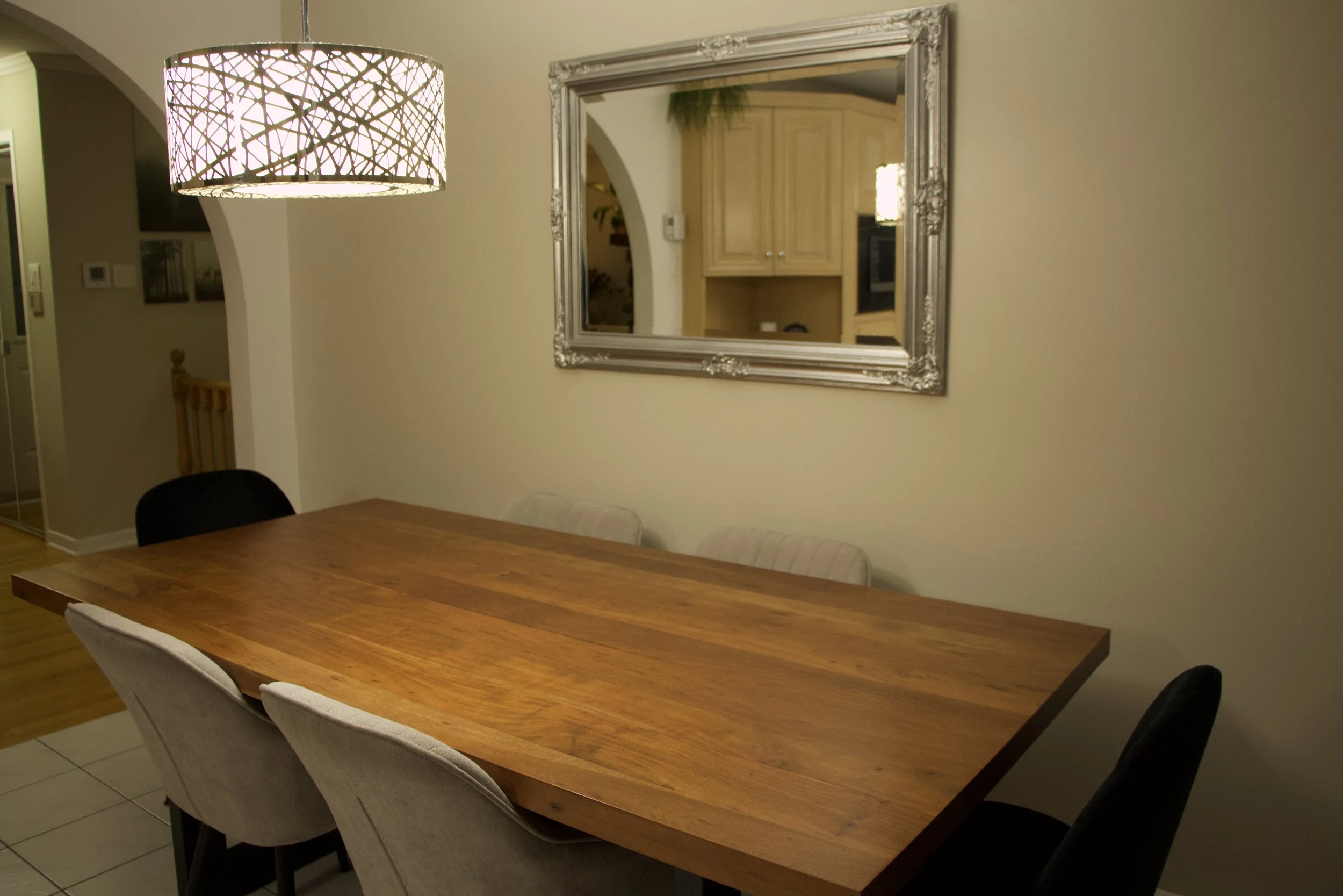 Interior view of a dining area with a wooden table, upholstered chairs, a decorative mirror on the wall, ceiling light fixture, and a kitchen with light wood cabinets reflected in the mirror.