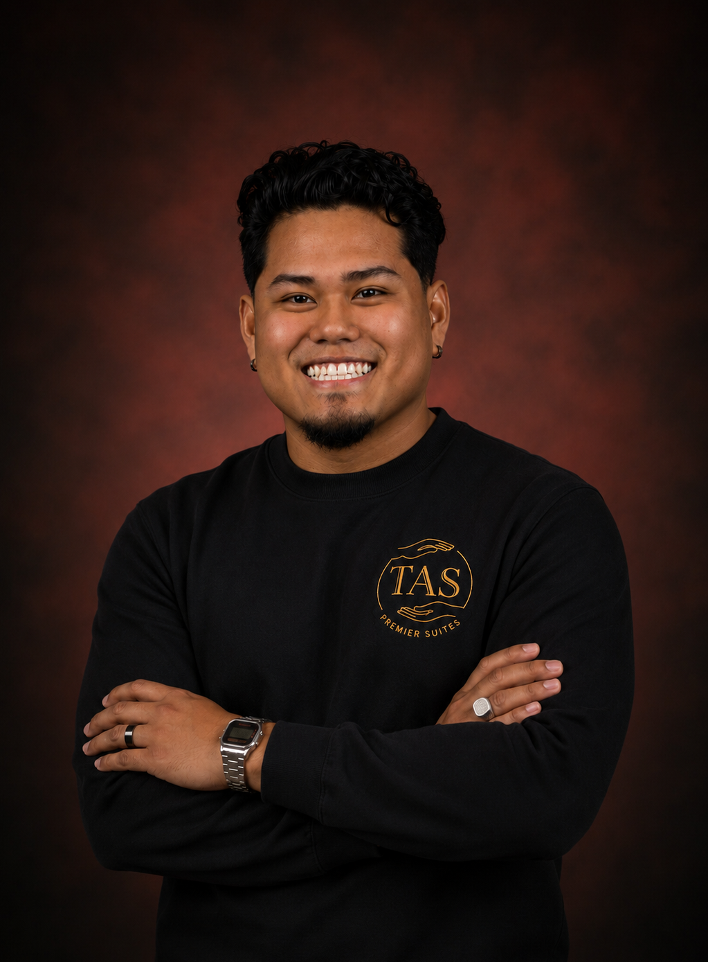A young man with short curly black hair and earrings, smiling with arms crossed, wearing a black sweatshirt with a gold logo that reads 'TAS Premier Suites'.