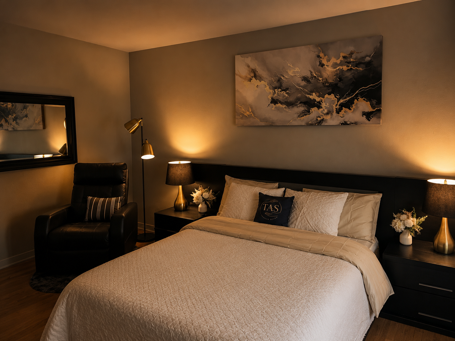 A cozy bedroom with a neatly made bed, black leather armchair, mirror, and warm lighting from lamps, with a large abstract painting on the wall.