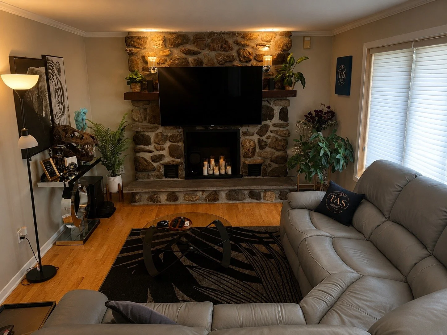 Living room with a gray leather sofa, a black coffee table, a stone fireplace with candles, a wall-mounted flat-screen TV, plants, a floor lamp, and window blinds.