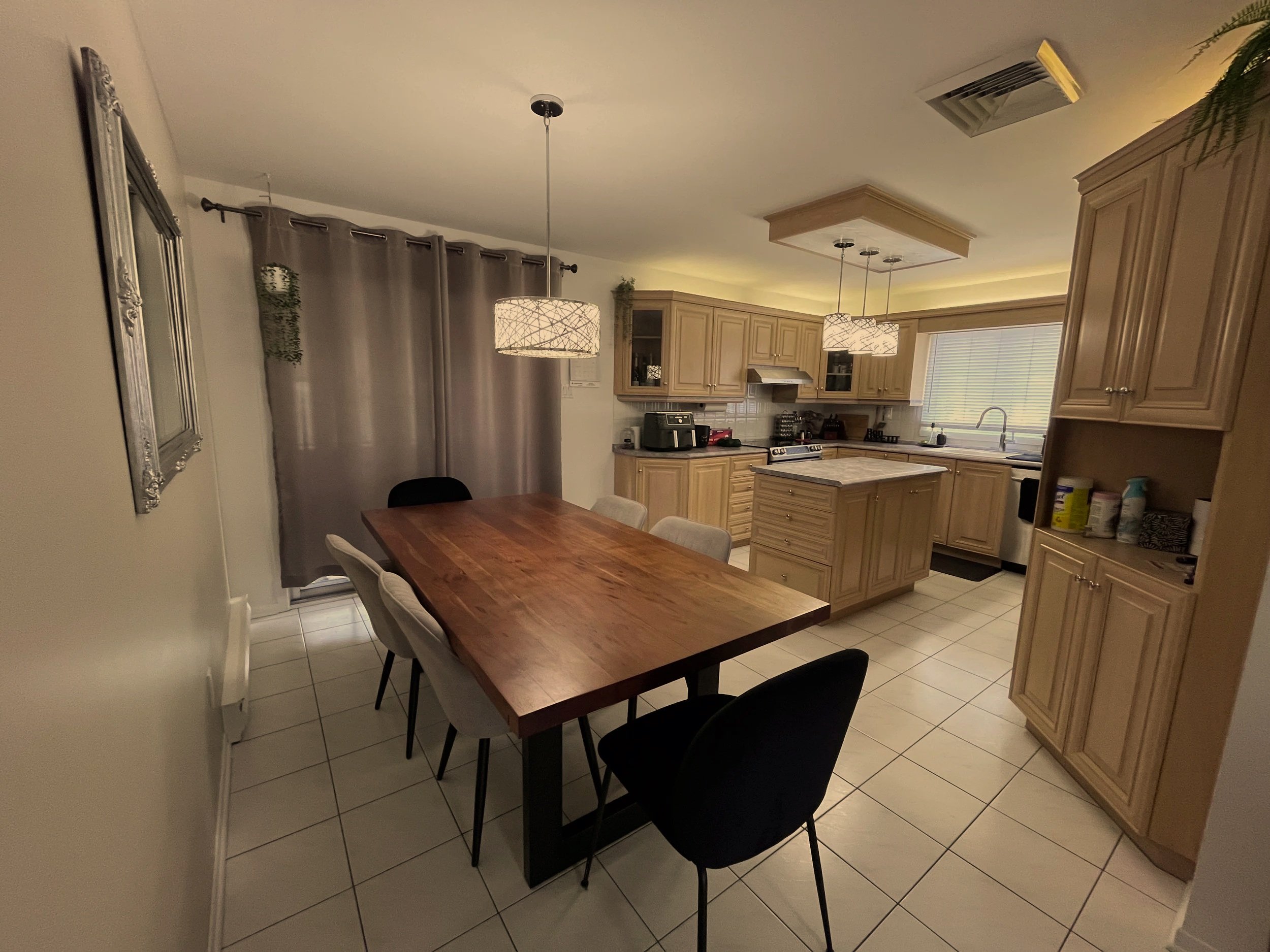 A dining area with a wooden table and black chairs, adjacent to a kitchen with light wood cabinets, a tiled backsplash, and a window with blinds. Pendant lights hang from the ceiling, and a gray curtain covers a door.
