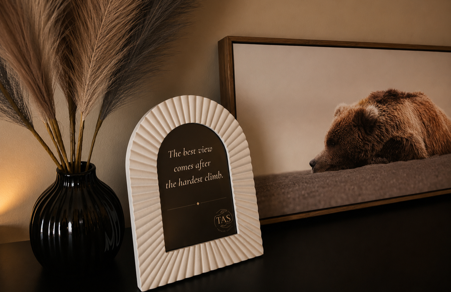 A black vase with tall, dried pampas grass, a framed quote print that reads 'The best view comes after the hardest climb,' and a framed photo of a bear lying down.