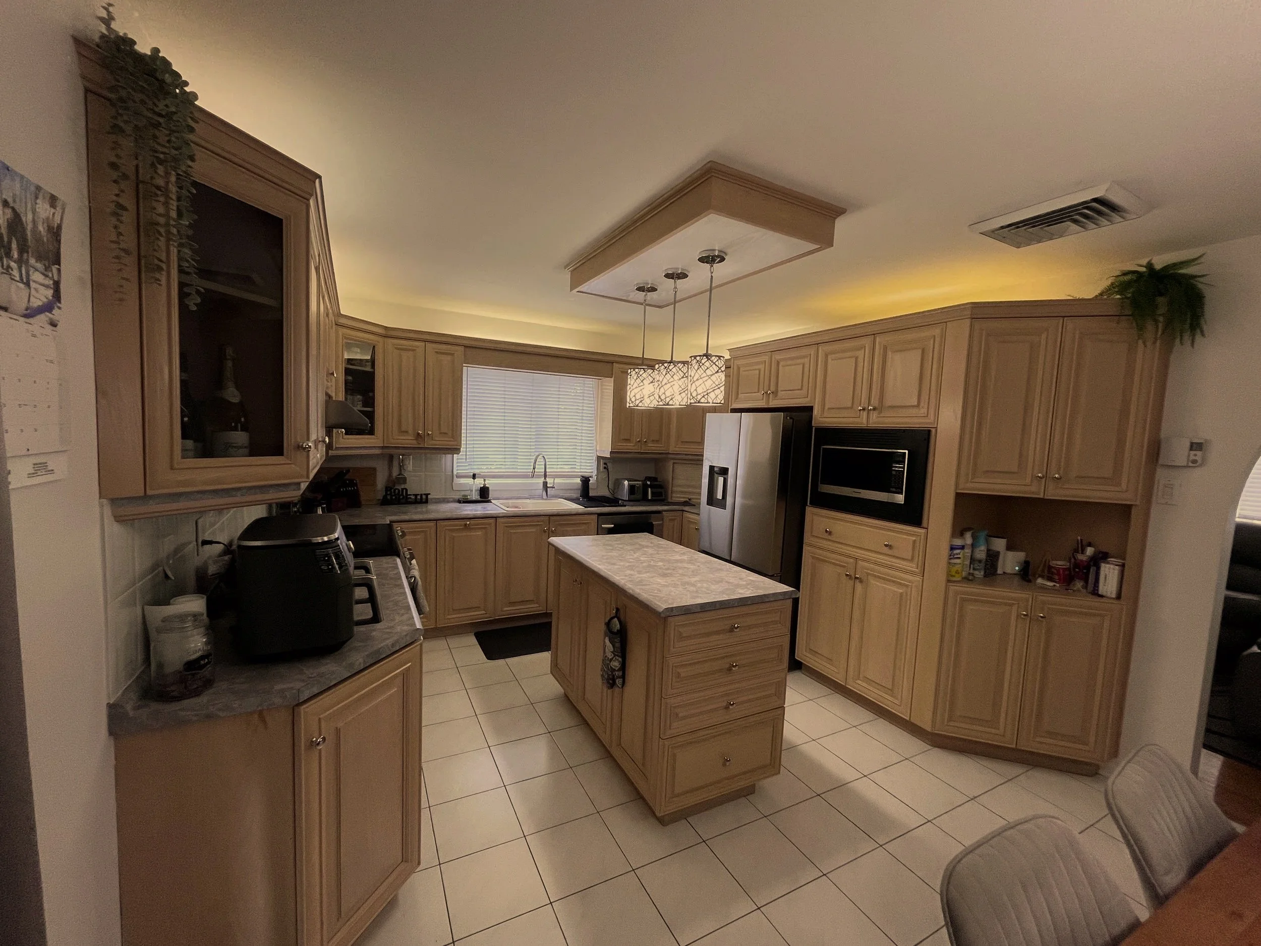 Kitchen with wooden cabinets, tile floor, and a window with blinds. Pendant lights over the kitchen island, a microwave oven, and a refrigerator are visible. Various kitchen appliances and items are on the countertops.