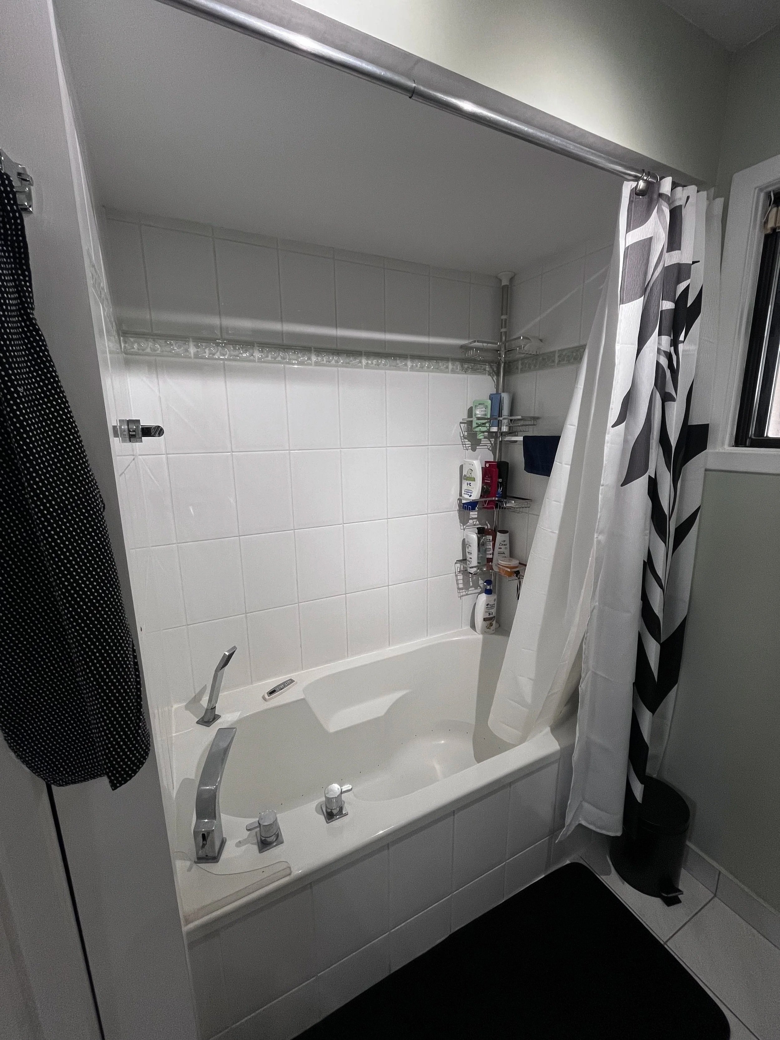 A white bathtub with silver fixtures in a bathroom, with a shower curtain partially drawn to the side, displaying a shelf with various toiletries and a window with a partially closed blind.