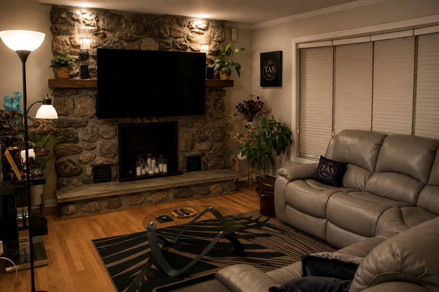 A cozy living room with a stone fireplace, mounted flat-screen TV, and beige leather sofas. Decor includes plants, candles, a floor lamp, and a rug with a geometric pattern.