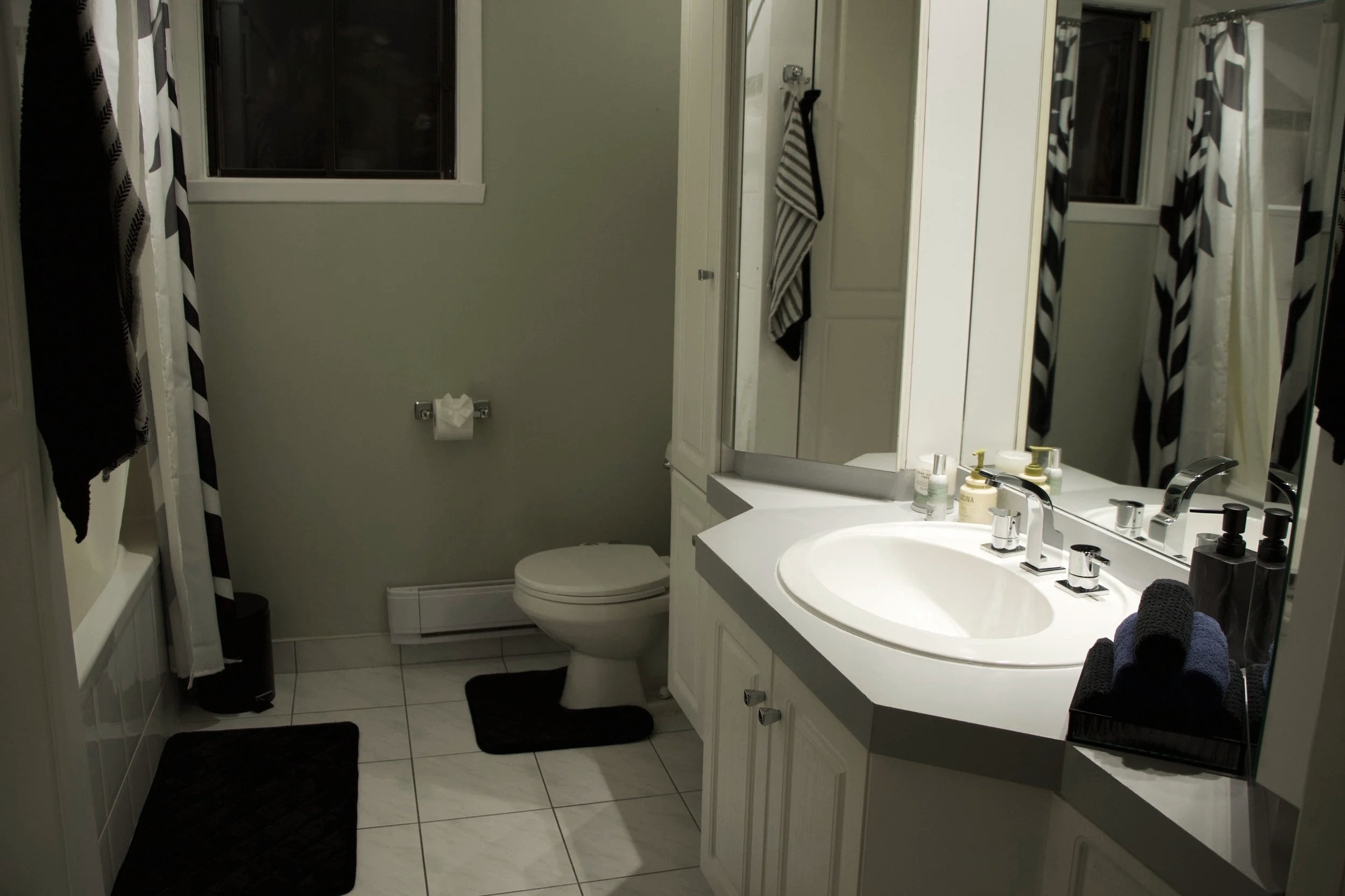 A small bathroom with a white vanity, oval sink, and mirrored cabinet. The vanity has various toiletries on it. To the left, curtains with black and white diagonal stripes are visible. There’s a toilet with a tissue roll on the wall above it. A window with a dark frame is on the wall. The floor has white tiles, and there are black mats on the floor.