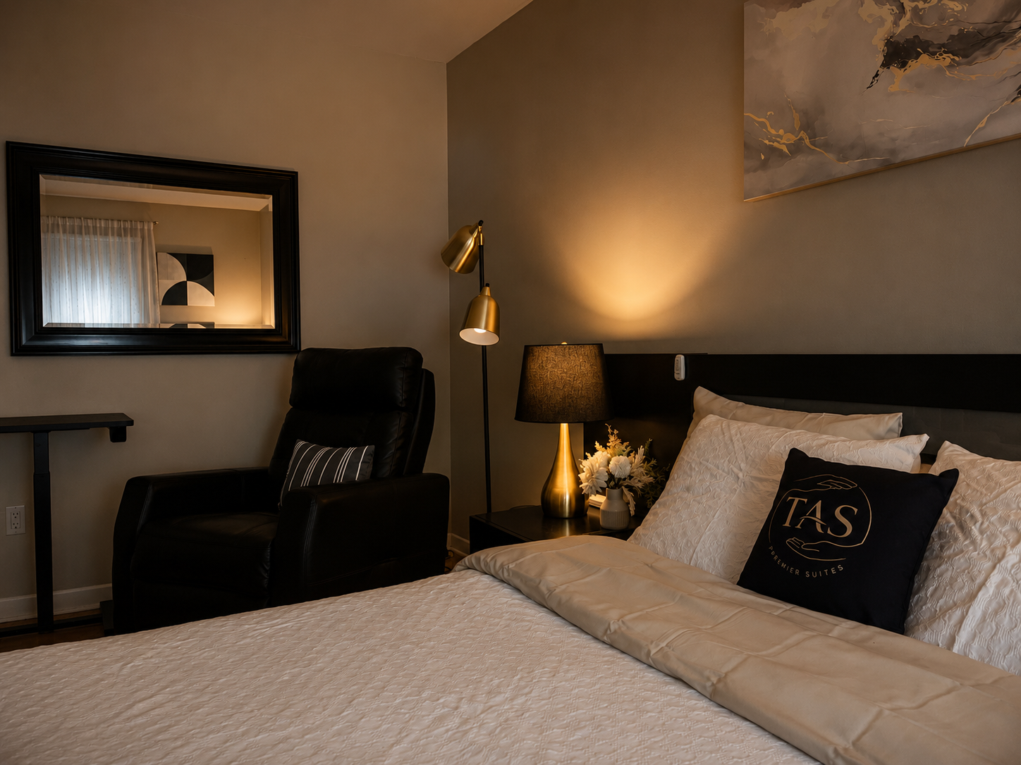A hotel room featuring a neatly made bed with white bedding, a black headboard, a decorative black pillow with gold lettering, a side table with a black lamp, a floor lamp with gold accents, a black leather armchair, a mirror on the wall, and a window with sheer curtains.
