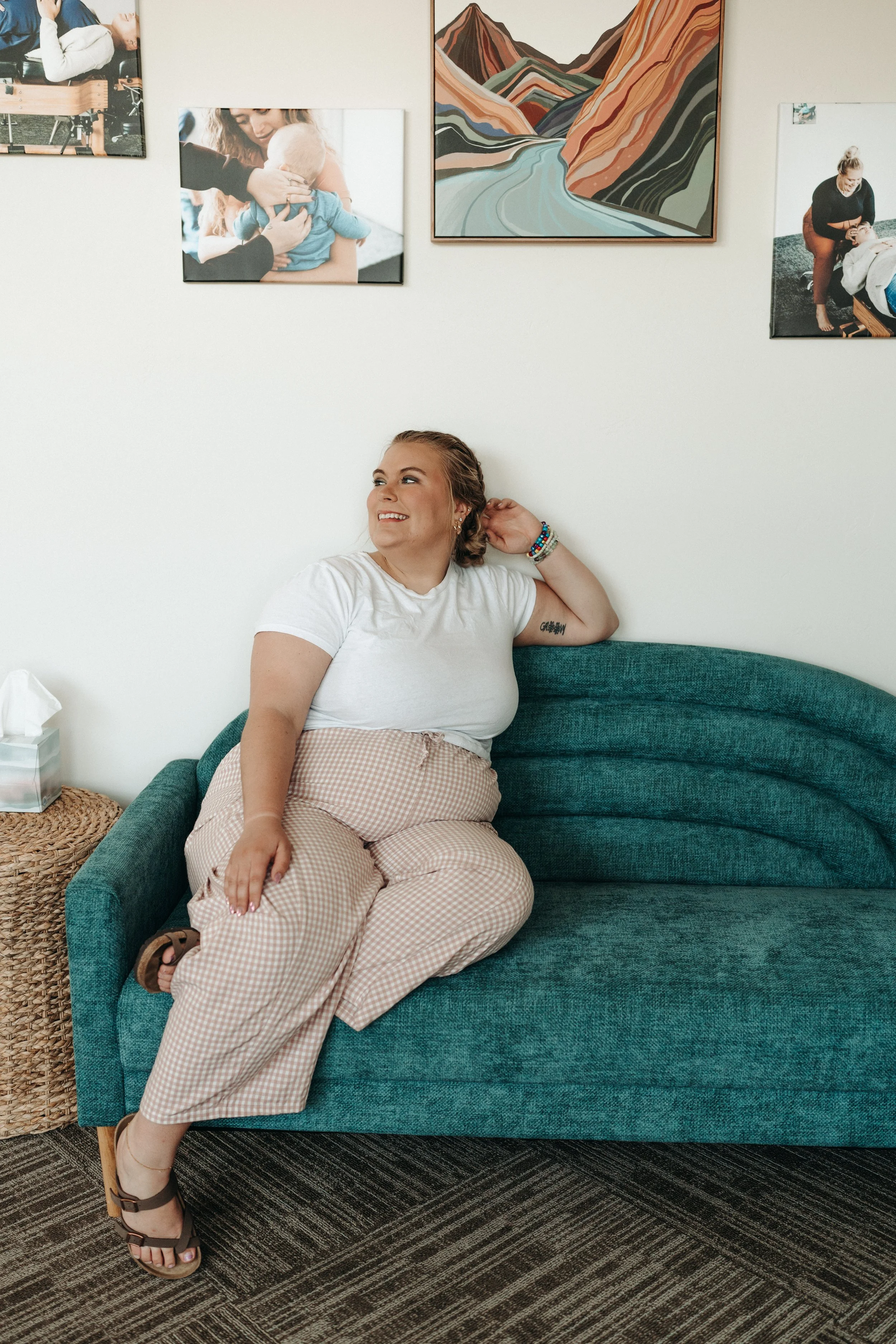A young woman with short hair and a tattoo on her arm sitting on a teal couch, smiling and looking up, wearing a white t-shirt, patterned pants, and sandals. The wall behind her has framed photos and a colorful mountain landscape painting.