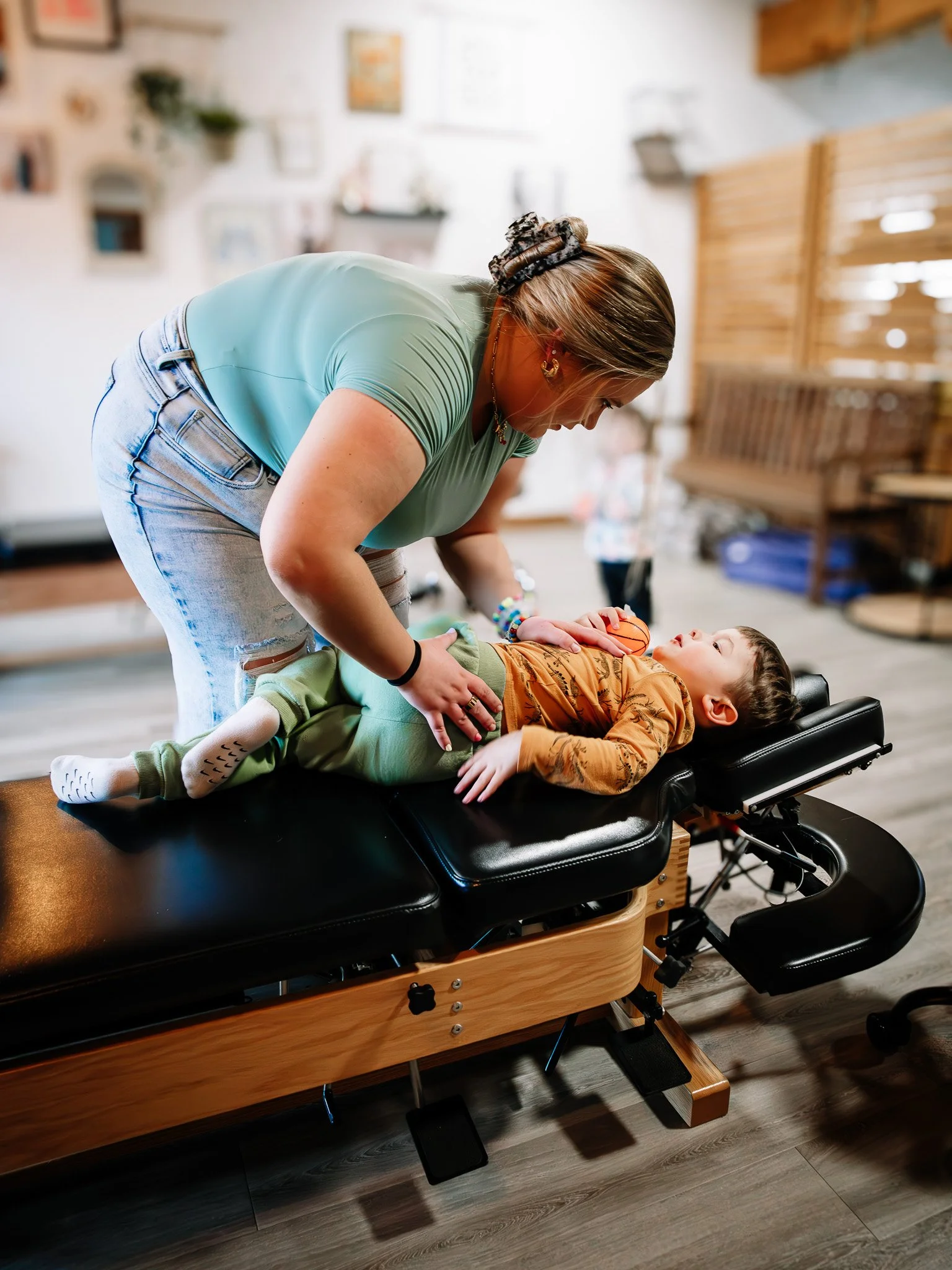 A woman with a scrunchie in her hair leaning over a young boy lying on a chiropractic or massage table in a wellness or therapy room, with wooden accents and framed artwork on the wall in the background.