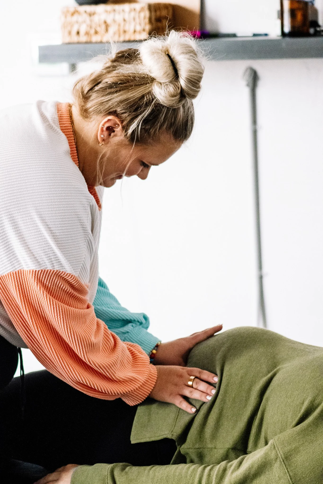 A woman is performing a massage on a person lying face down. The woman has blonde hair styled in a bun, and is wearing a white and orange ribbed sweater. The person receiving the massage is wearing green pants and a green jacket.