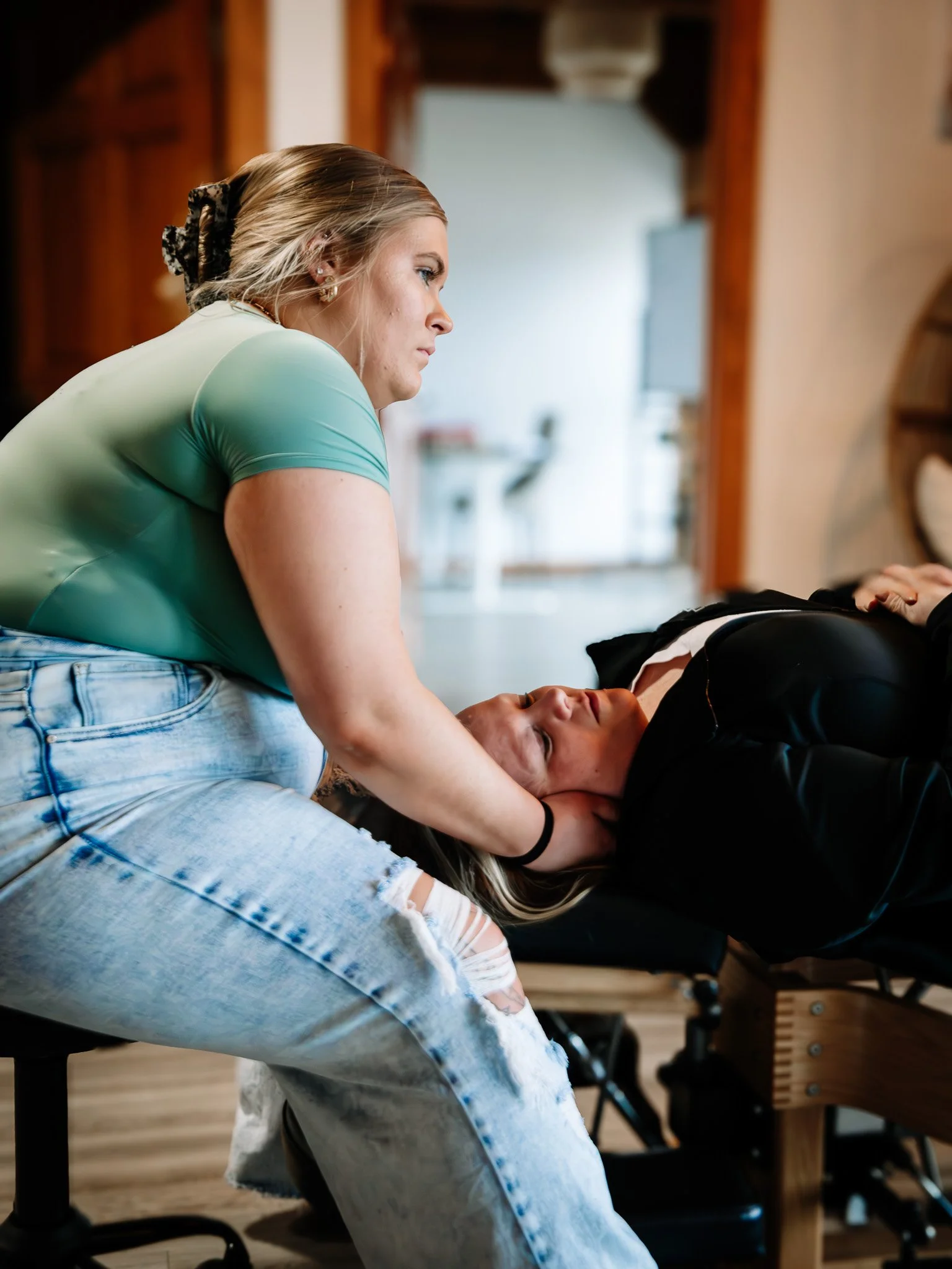 A woman with blonde hair in a leopard-print scrunchie wearing a light blue top and distressed jeans leaning over another woman with dark hair lying down.