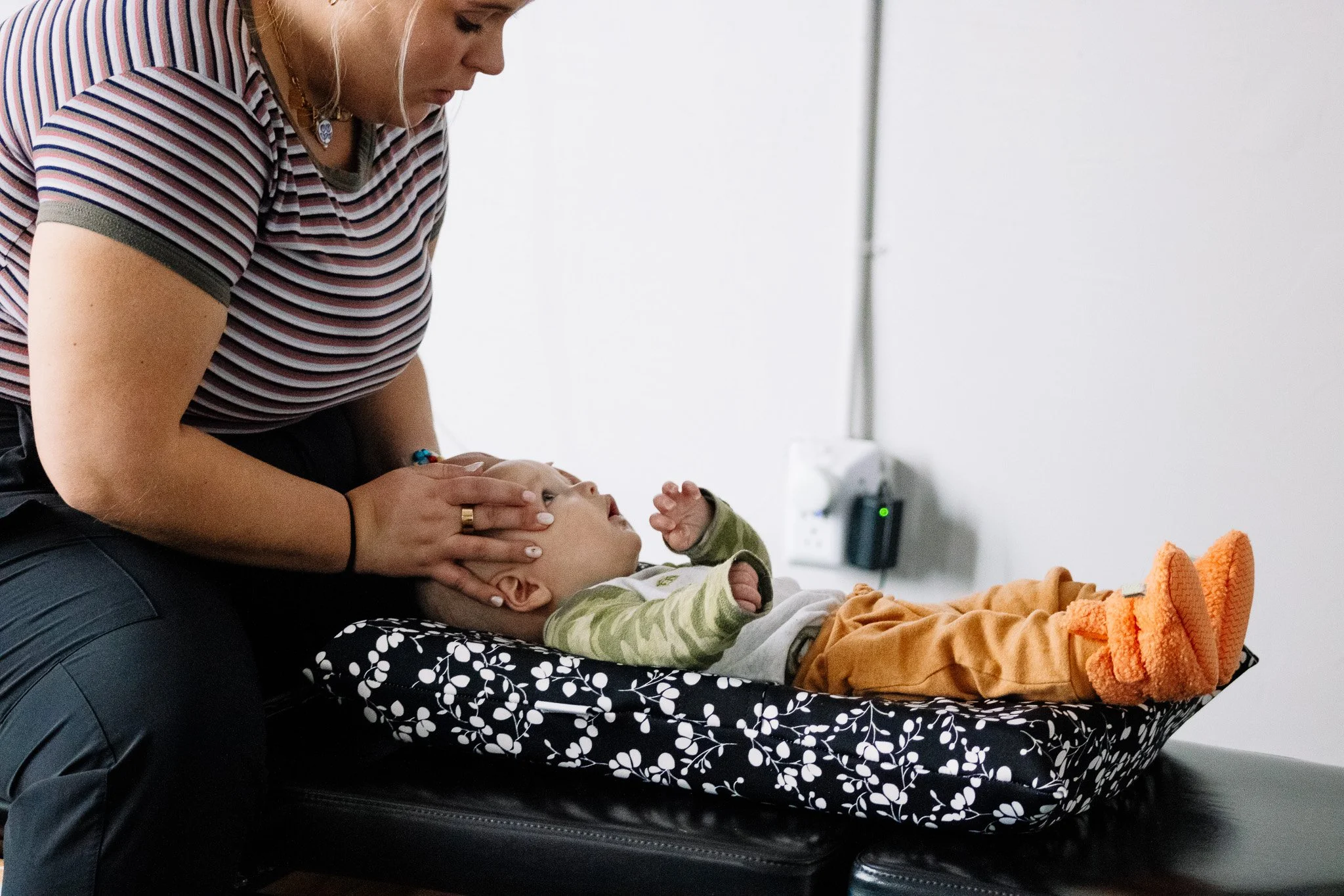 A woman in a striped shirt is gently holding the head of a young boy lying on a padded examination table, possibly during a medical or dental checkup.